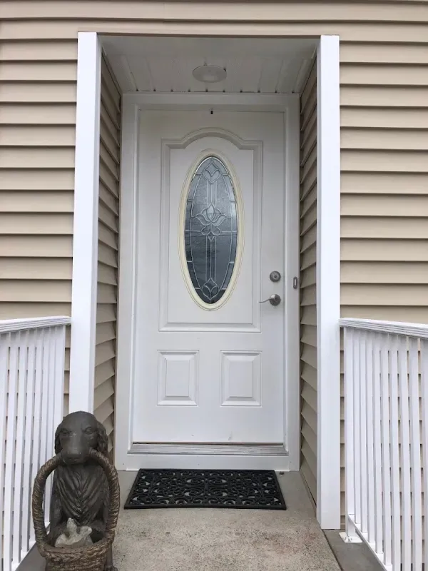 A white front door with an oval glass pane, framed by tan siding and white railings, with a dog-shaped doorstop on a mat.