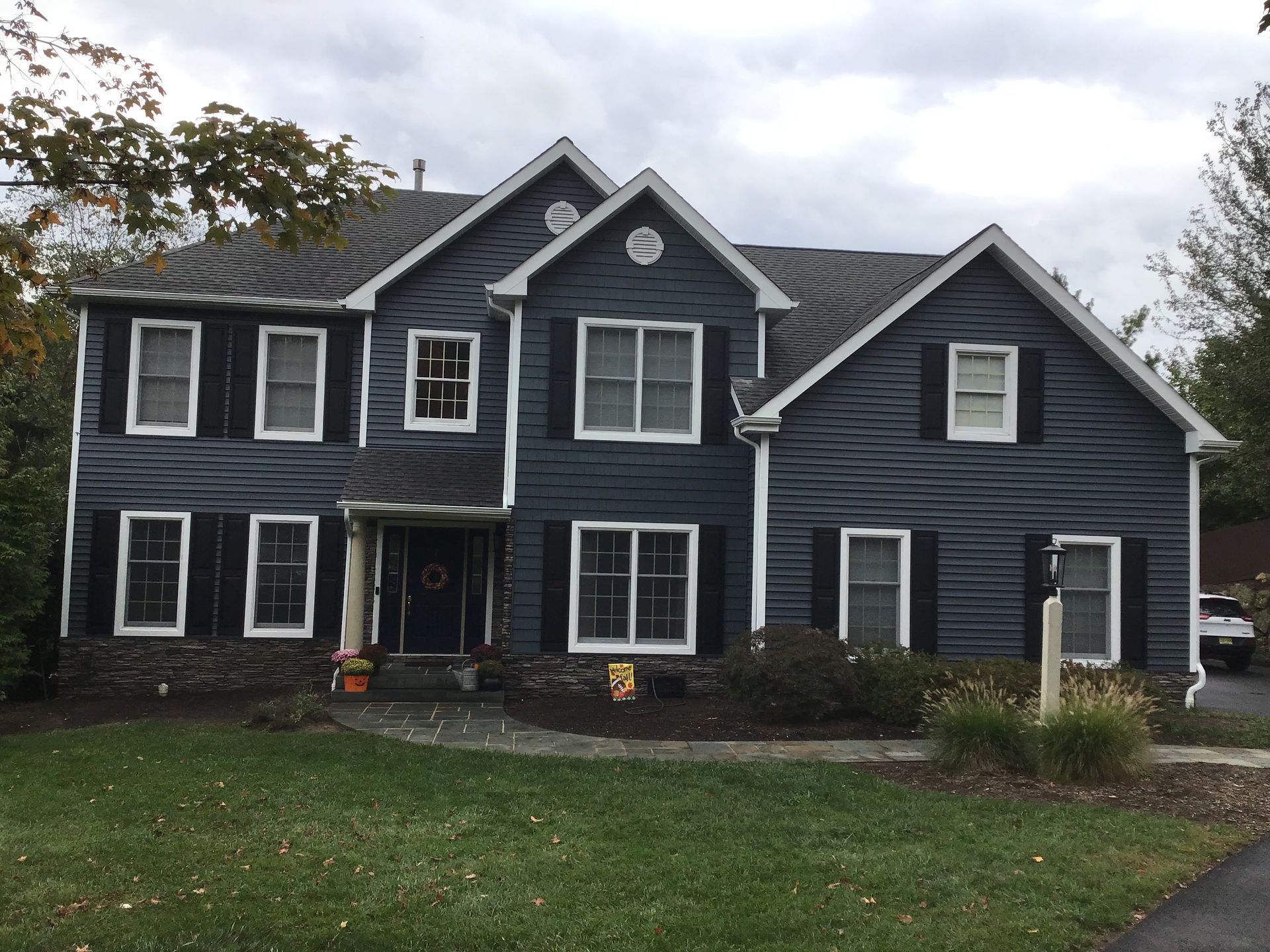 A two-story dark blue suburban home with white trim, black shutters, and a stone facade on a cloudy day.