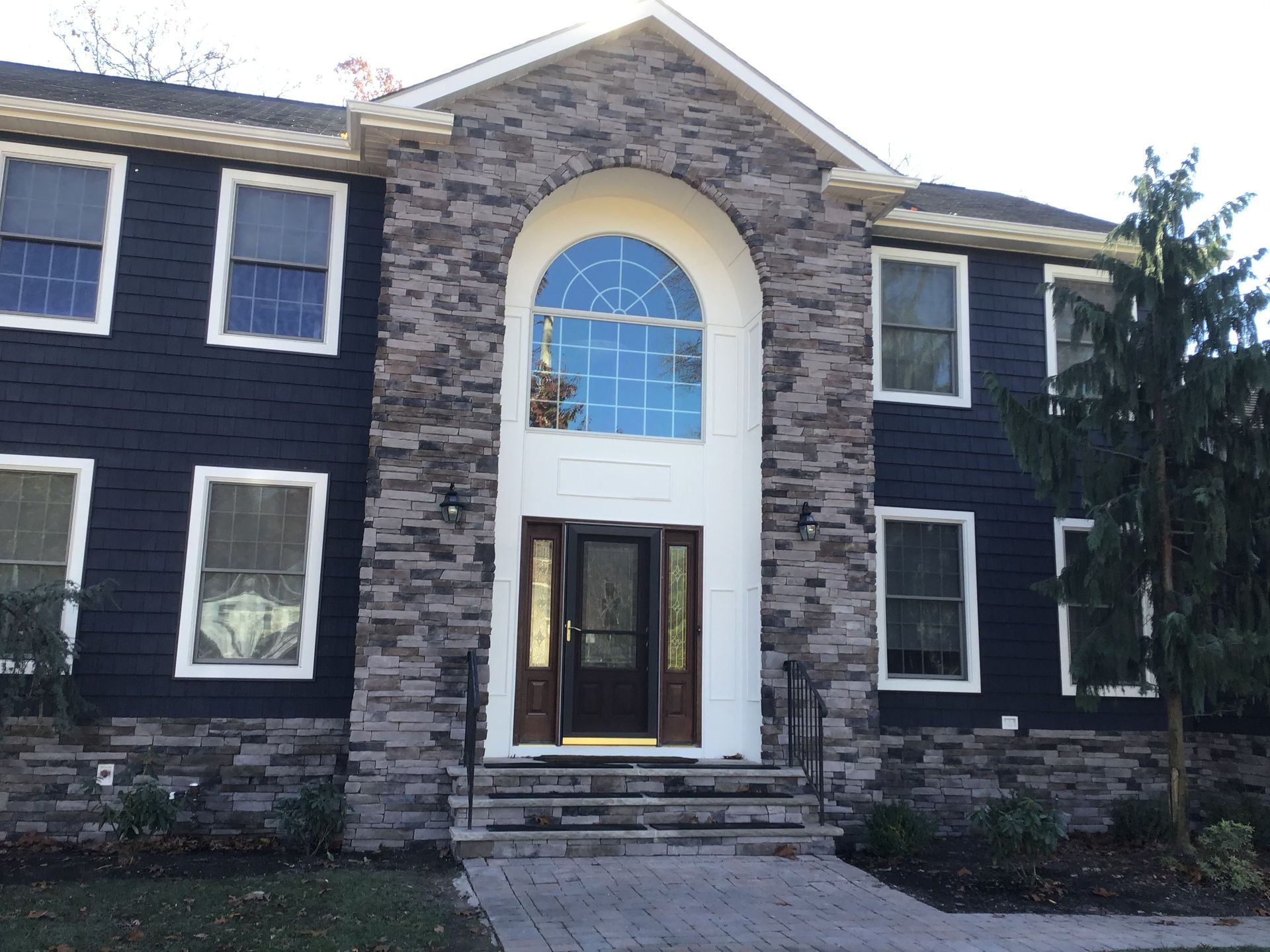 A two-story house with dark blue siding, a stone facade entryway, a tall arched window, and a paved walkway.