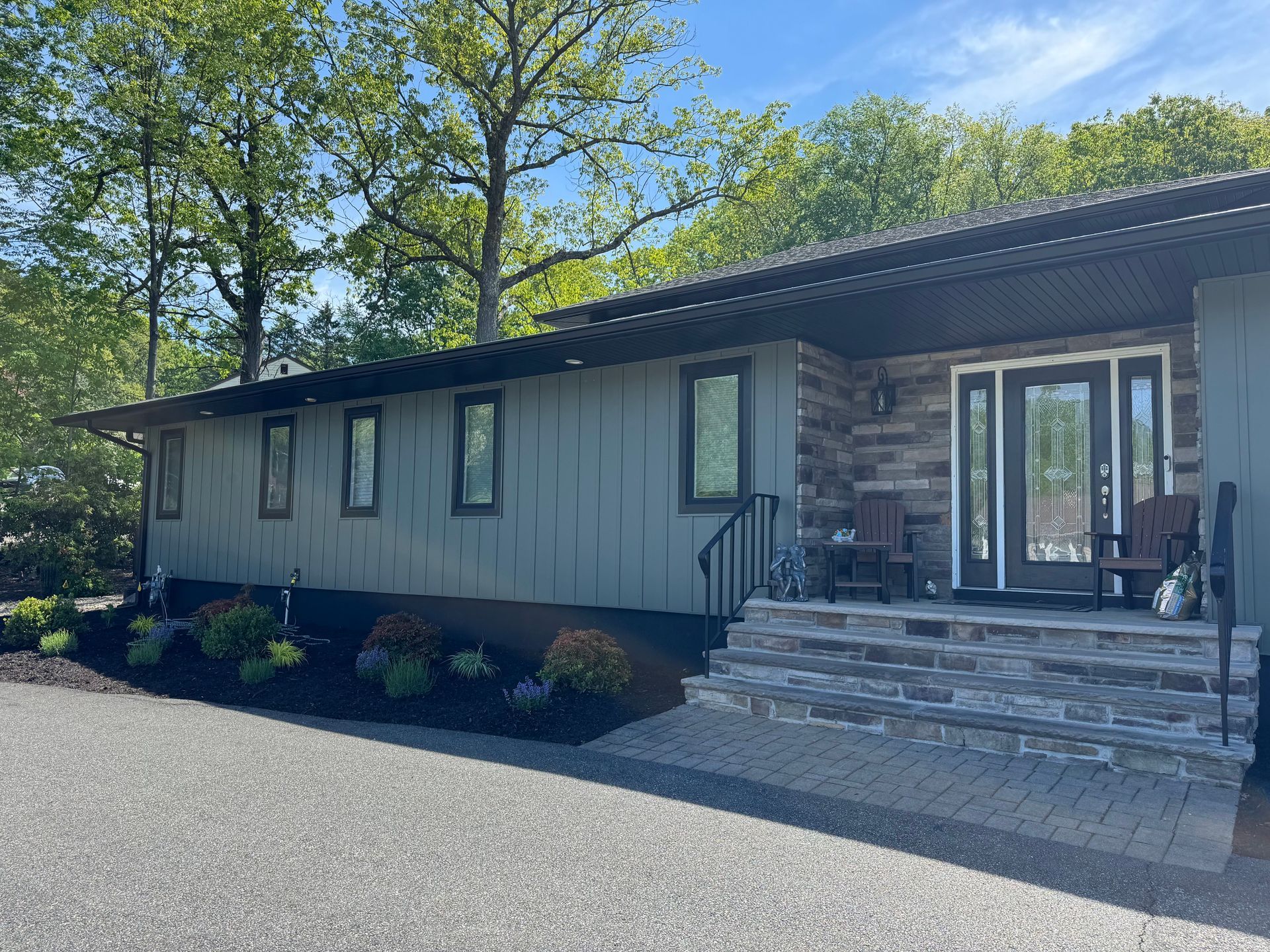 A single-story house with sage green vertical siding, a stone entryway, and a stone front porch on a gravel driveway.