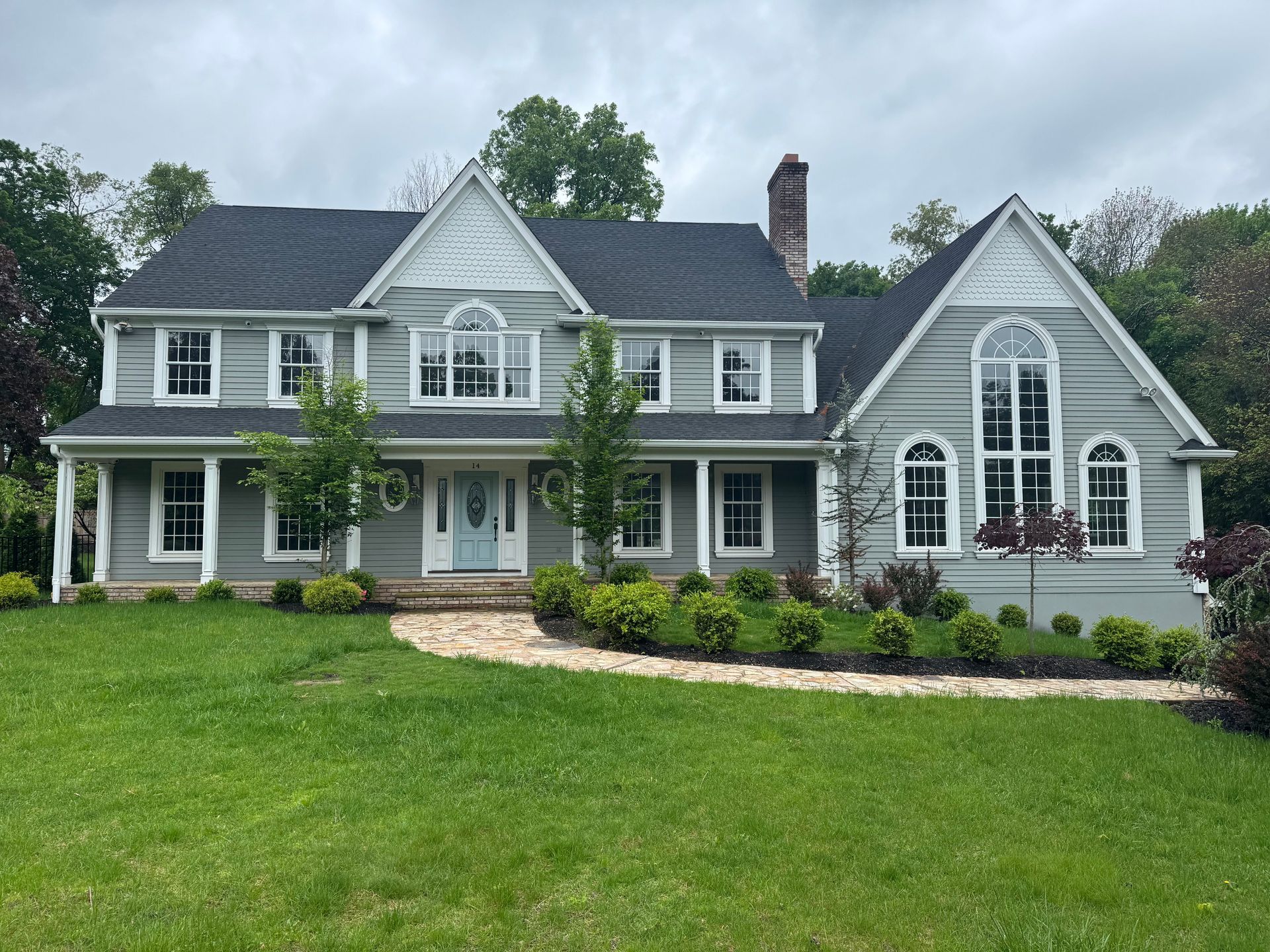 Two-story light gray house with a stone pathway, a front porch, and black shingled roof under a cloudy sky.