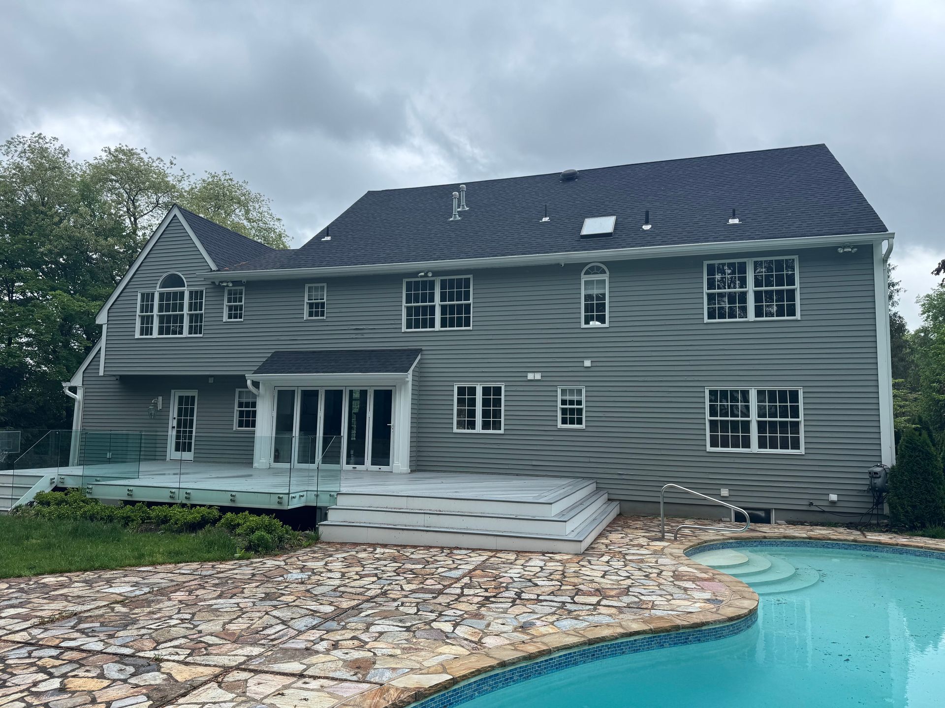 A two-story gray house with a backyard deck, stone patio, and swimming pool under a cloudy sky.