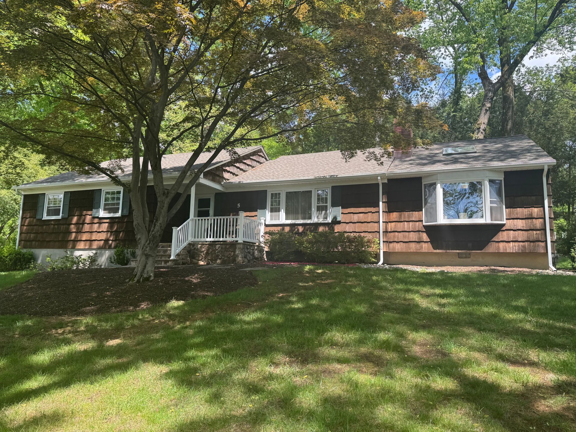 A one-story brown shingled house with a white front porch, large bay window, and a big tree in the front yard.