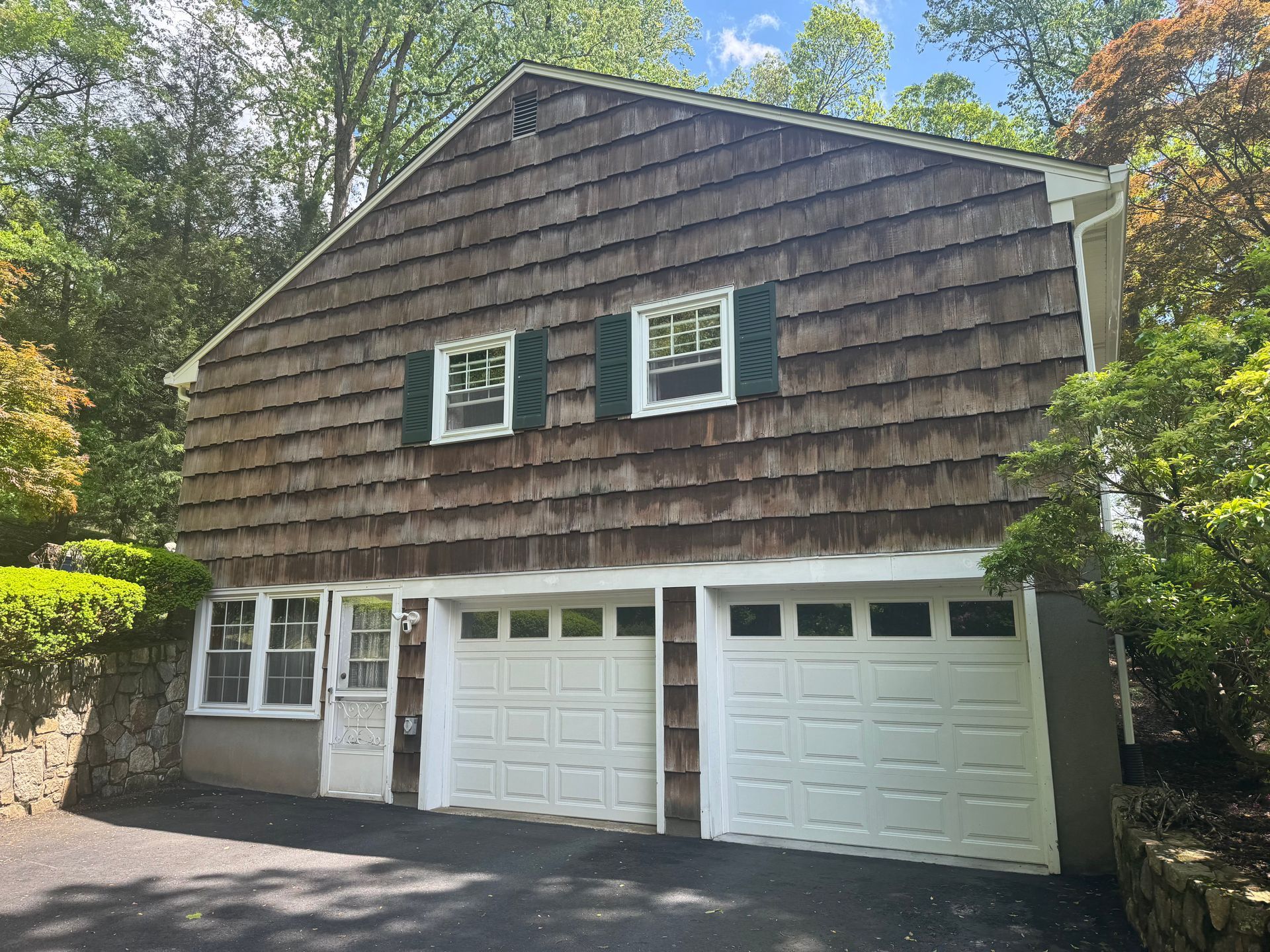 A two-story garage with brown wood shingle siding, two white garage doors, and two windows with green shutters.