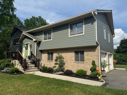 A two-story house with green, textured siding above a tan brick ground floor, featuring a front porch and gravel driveway.