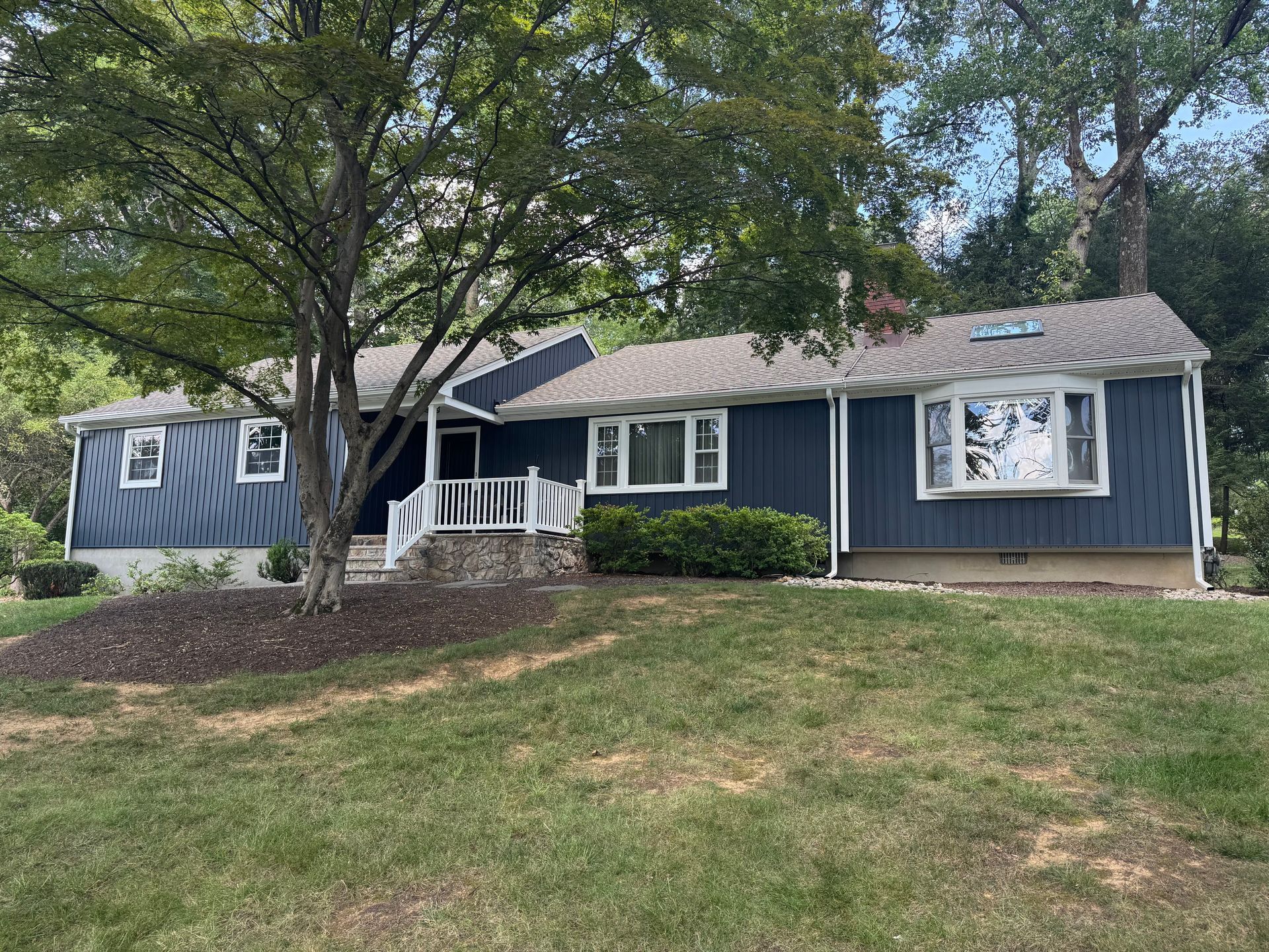 A dark blue ranch-style house with white trim, a bay window, and a small front porch, set on a green lawn with a large tree.