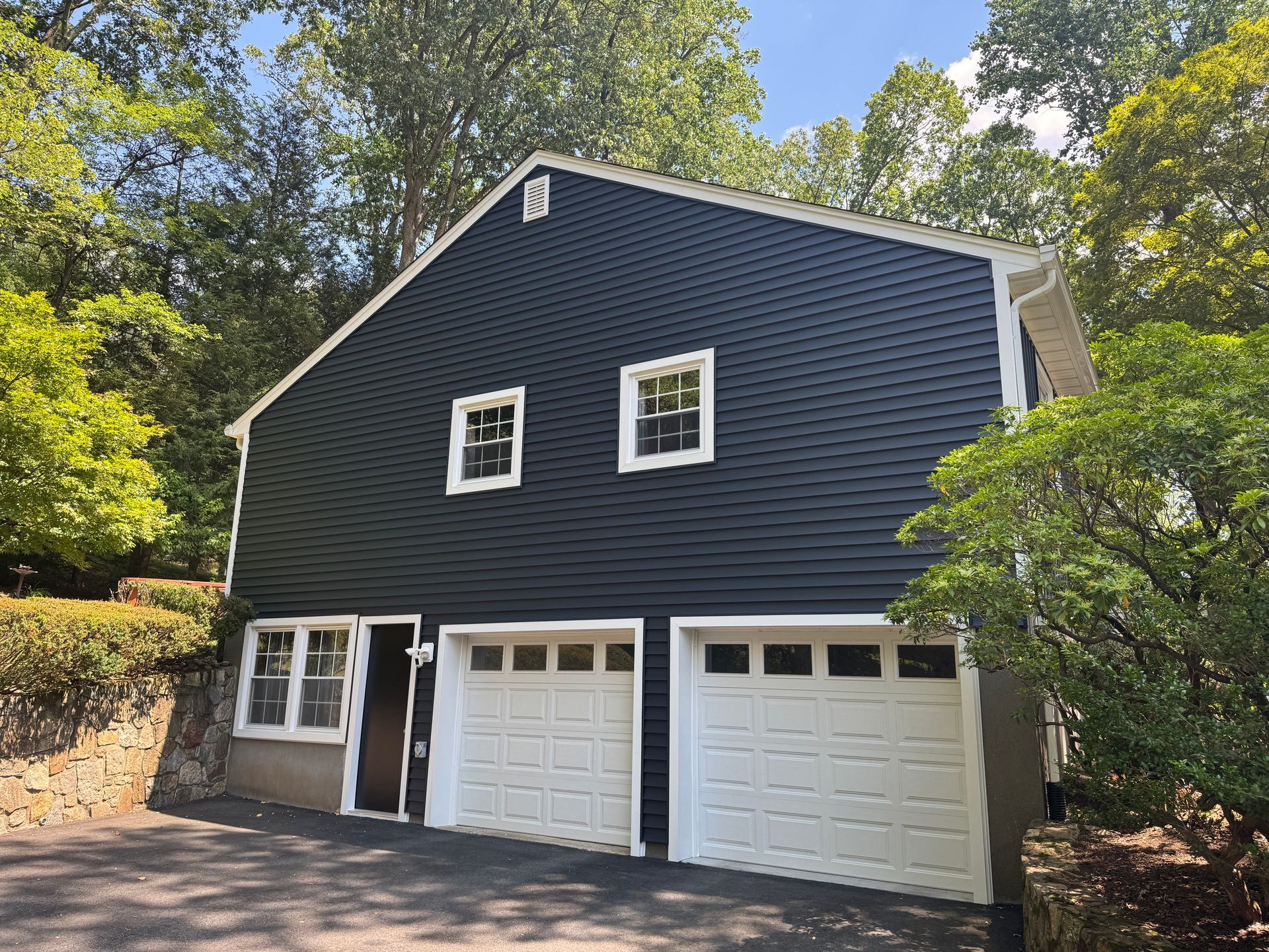 A two-story dark blue building with a two-car garage, white trim, and a small side entrance, set among lush trees.