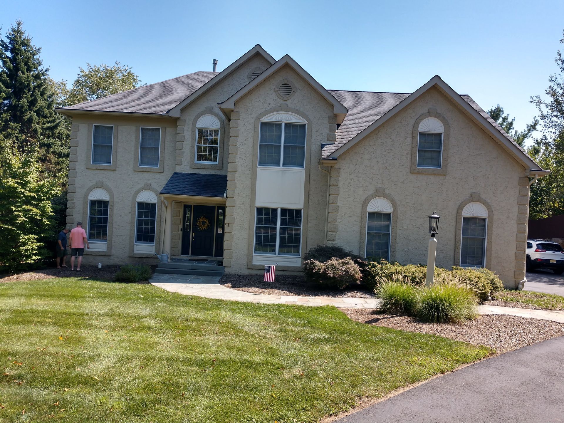A two-story tan brick house with a front porch, large windows, and a manicured lawn under a clear blue sky.