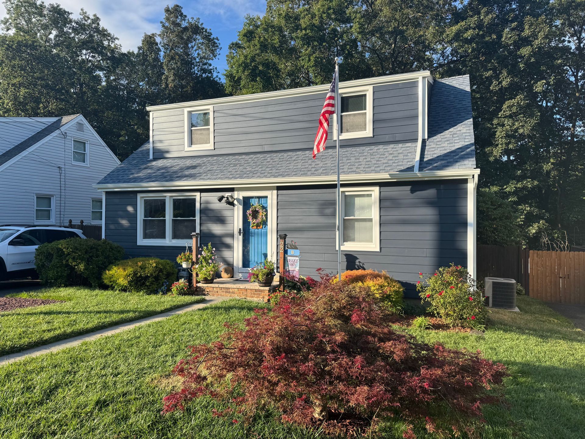 A two-story, blue-sided home with a dark shingled roof, a bright blue front door, and a flagpole in the front yard.
