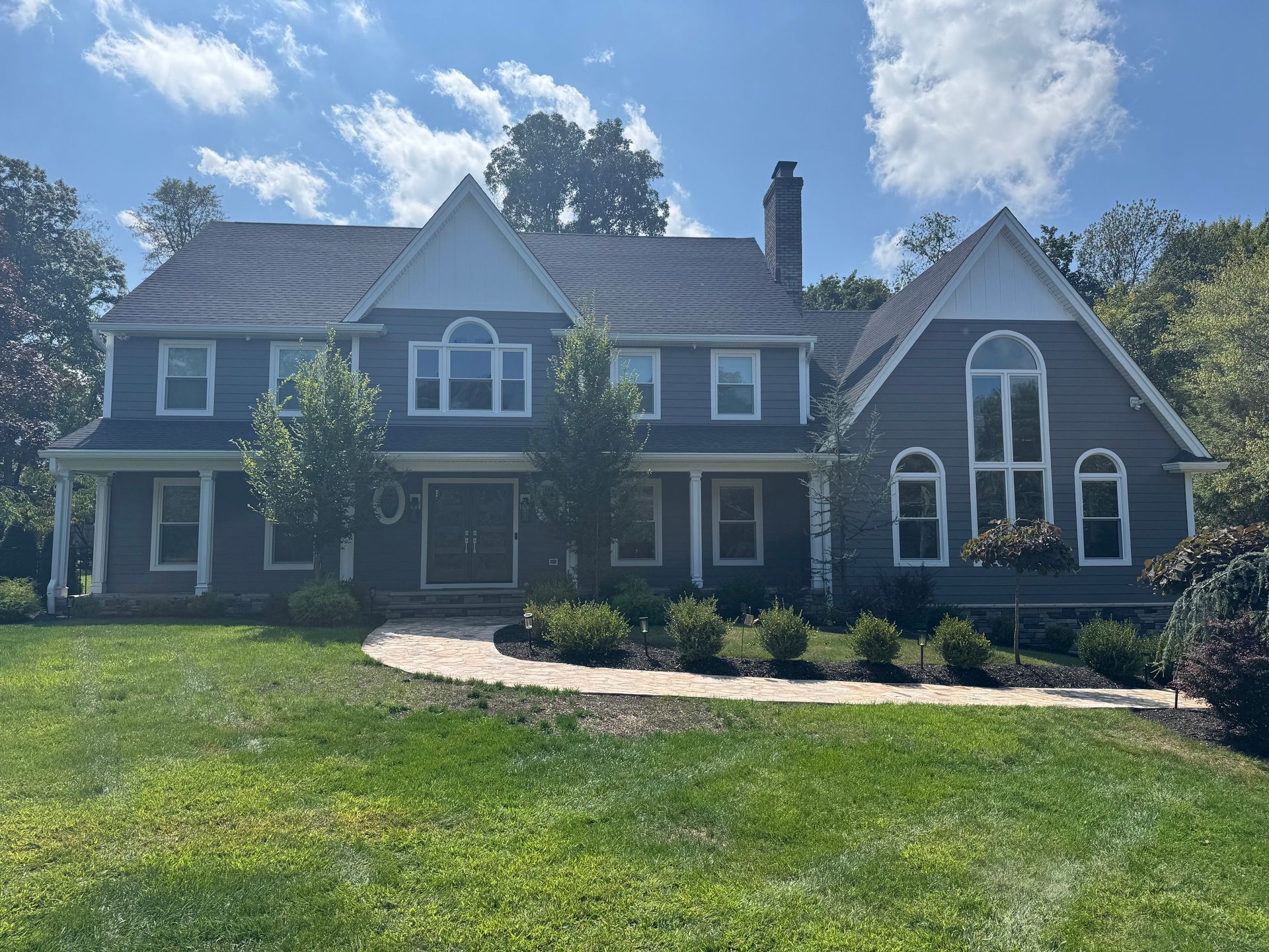 A large, two-story dark blue house with a white trim, a stone walkway, and a landscaped yard under a sunny blue sky.