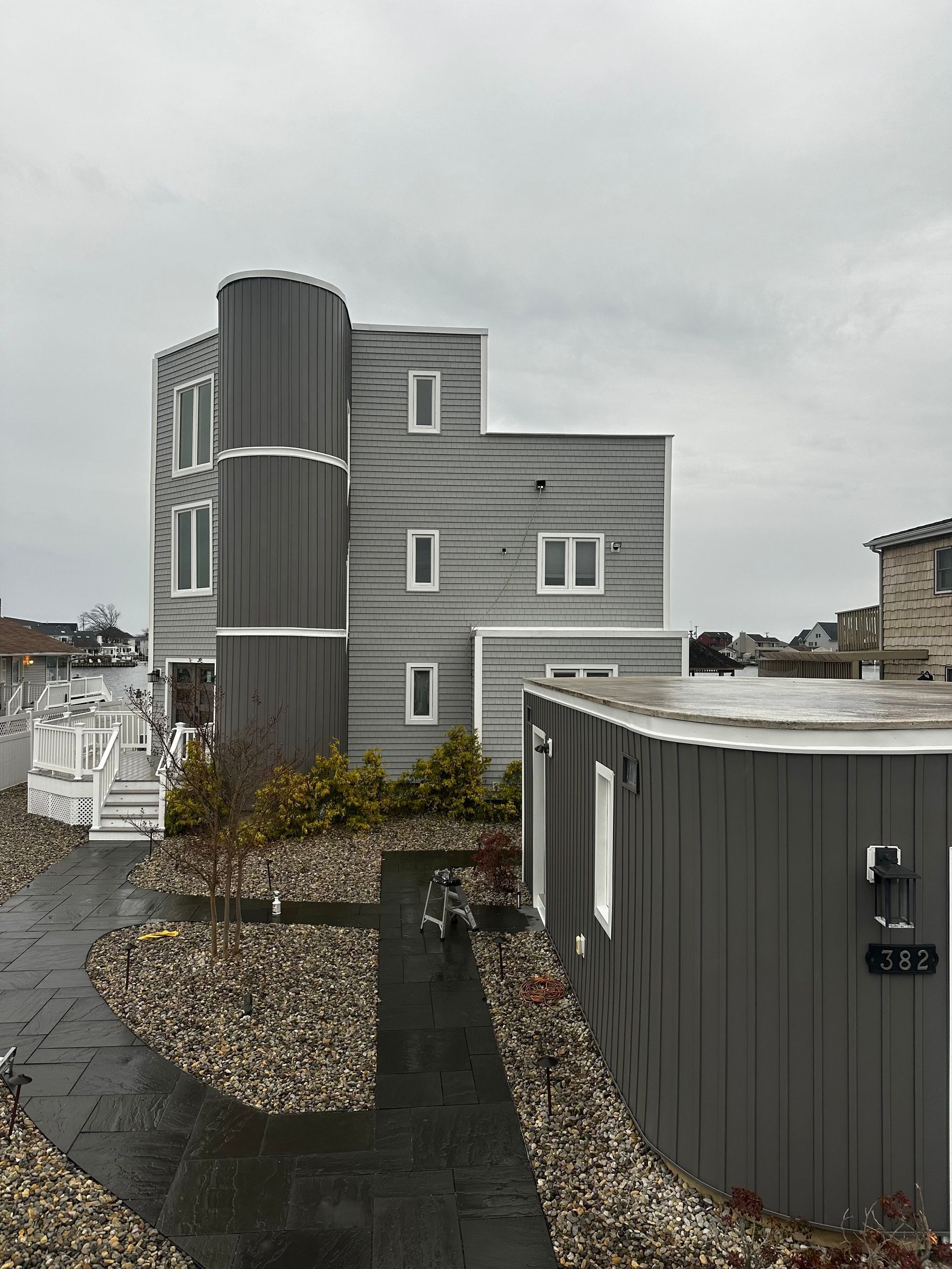 A modern, multi-story house with horizontal gray siding and a dark cylindrical tower section, next to a matching shed.