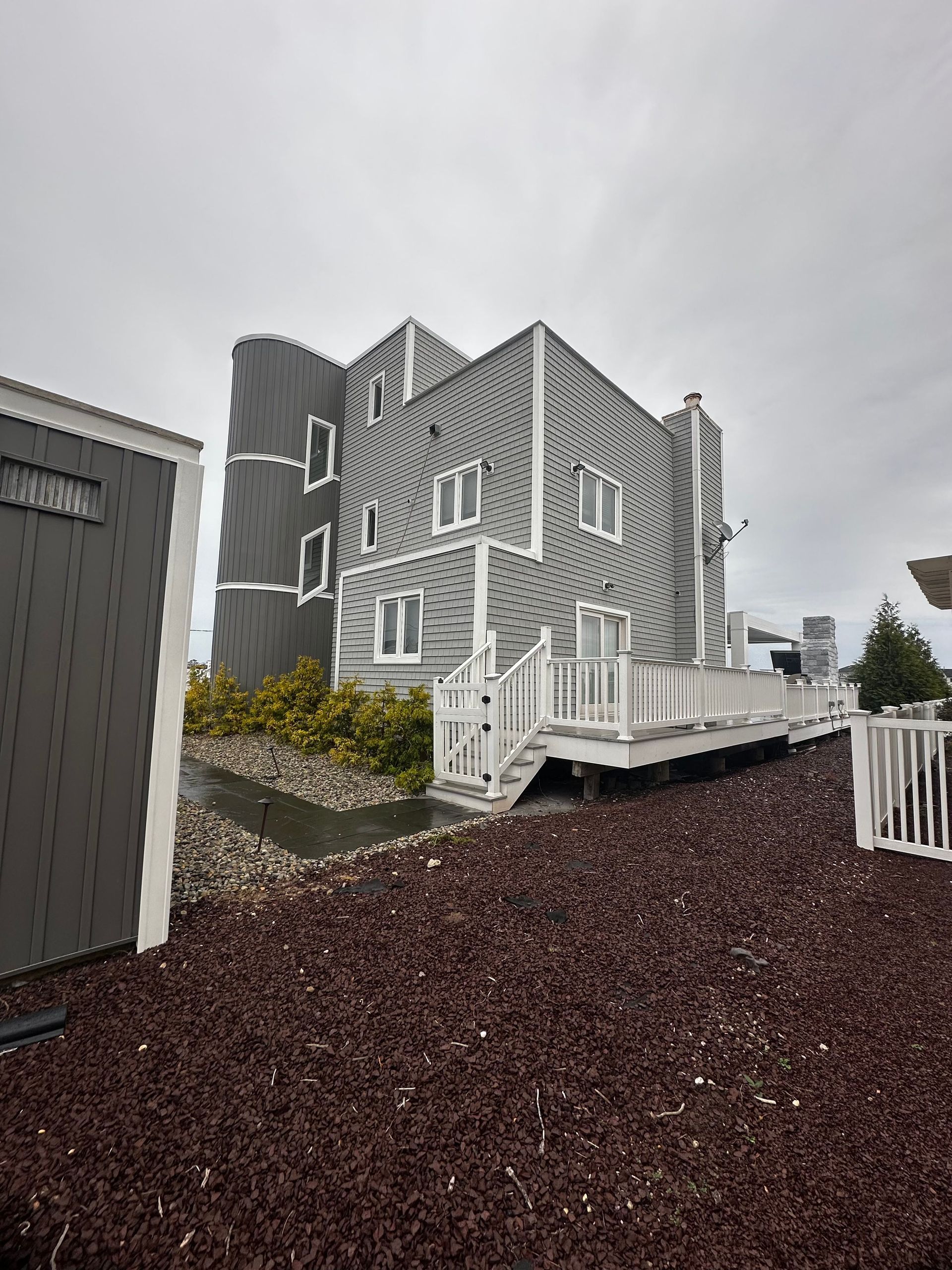 A three-story gray, shingled beach house with a white deck and a cylindrical tower feature, set against a cloudy sky.
