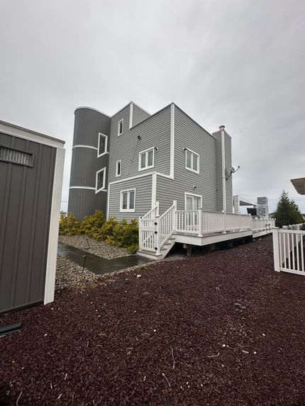 A gray, multi-story house with a rounded turret, white trim, and an attached deck over a dark gravel yard under a gray sky.