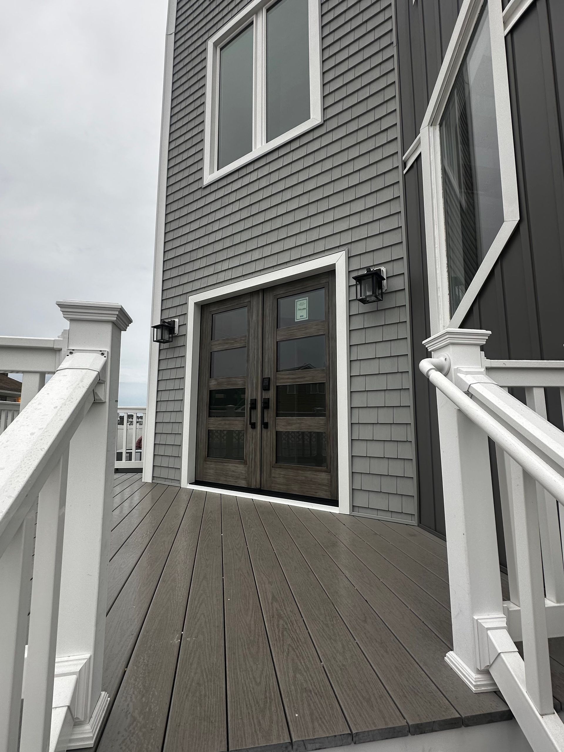 Gray shingled house exterior featuring a wooden double door entrance, a composite deck, and white railings.
