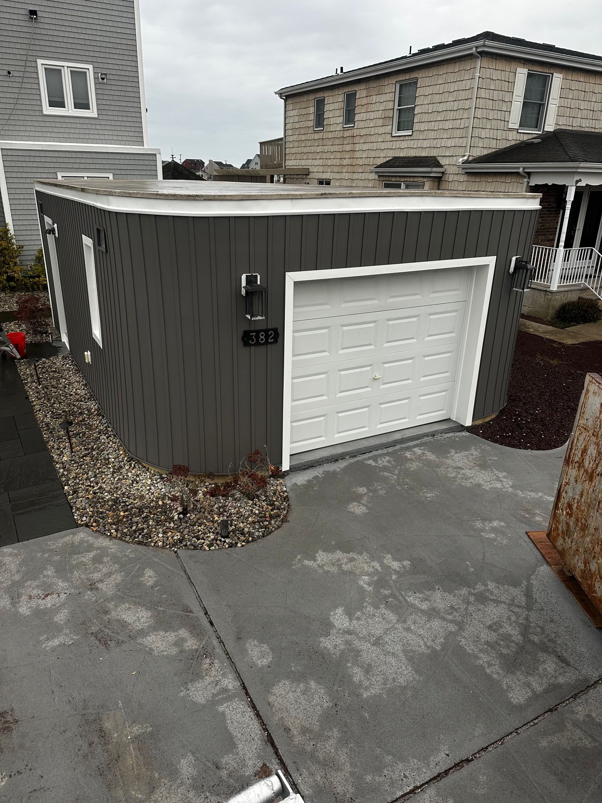 A detached, gray-sided garage with a white door and trim stands next to a concrete driveway and pebble landscaping.