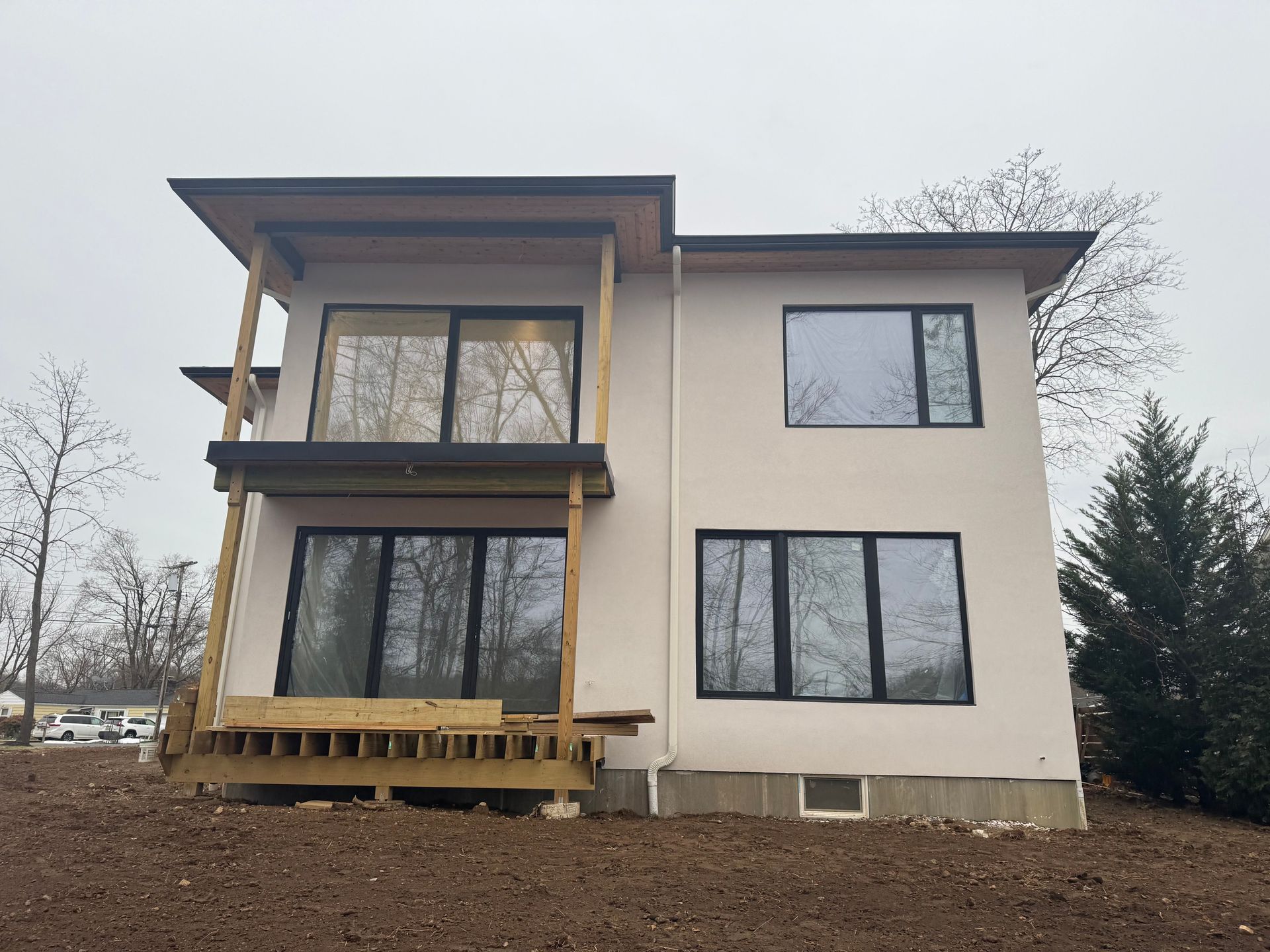 A two-story house with off-white stucco walls, black window frames, and an unfinished wooden deck and balcony.