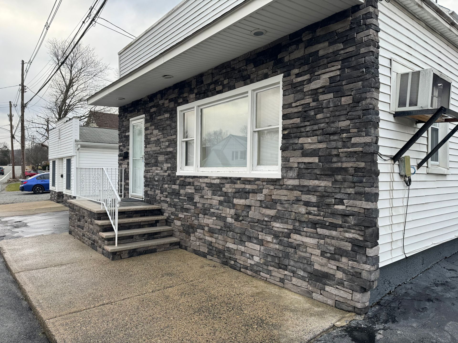 A stone-faced building with a concrete porch, short steps, and a white-framed window, next to a white siding structure.