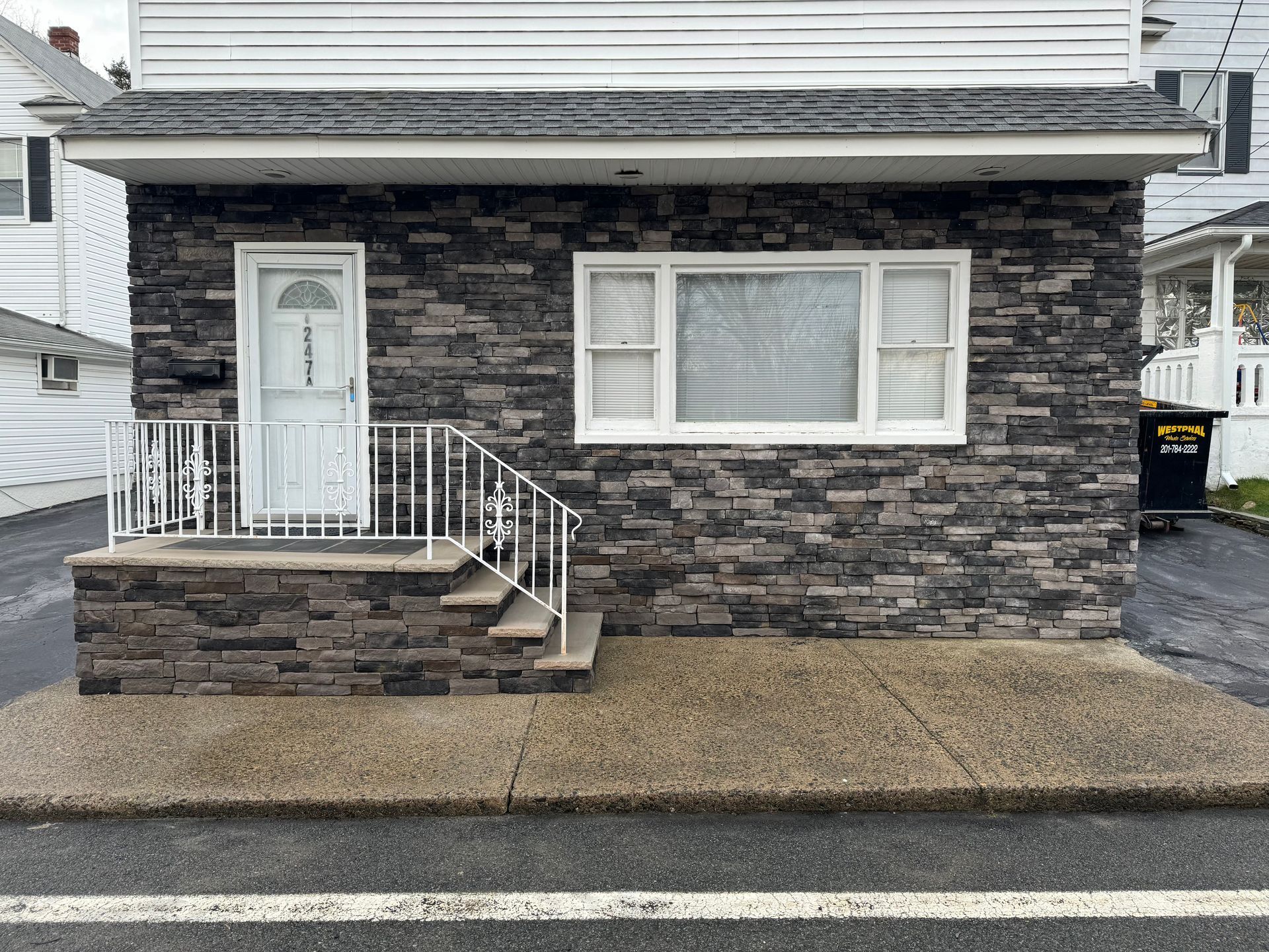 A small building front with a white door, a large window, white railing, and dark stone-textured siding.