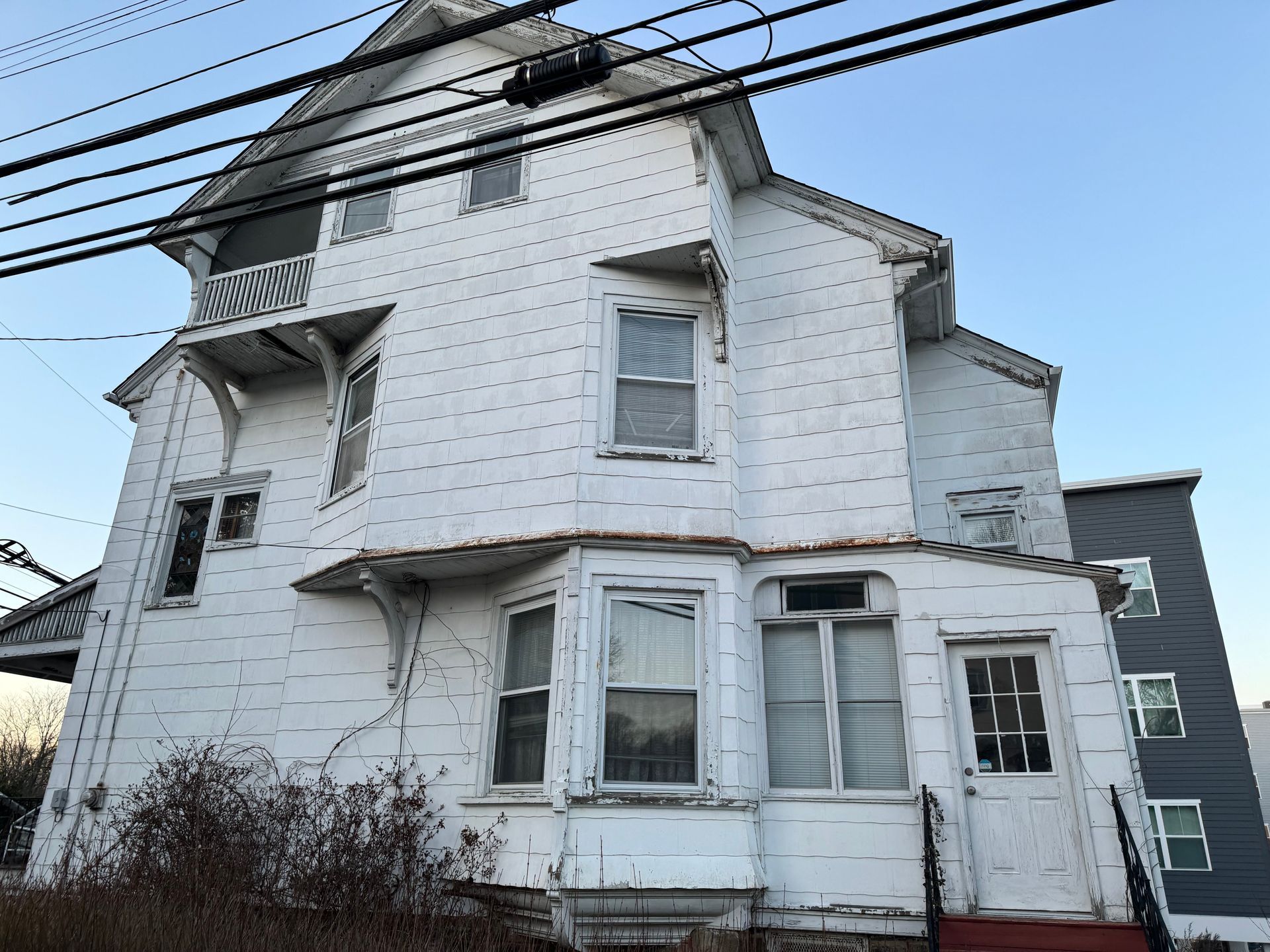 A weathered, white, three-story house with a bay window, porch, and exterior stairs, viewed from a low angle.