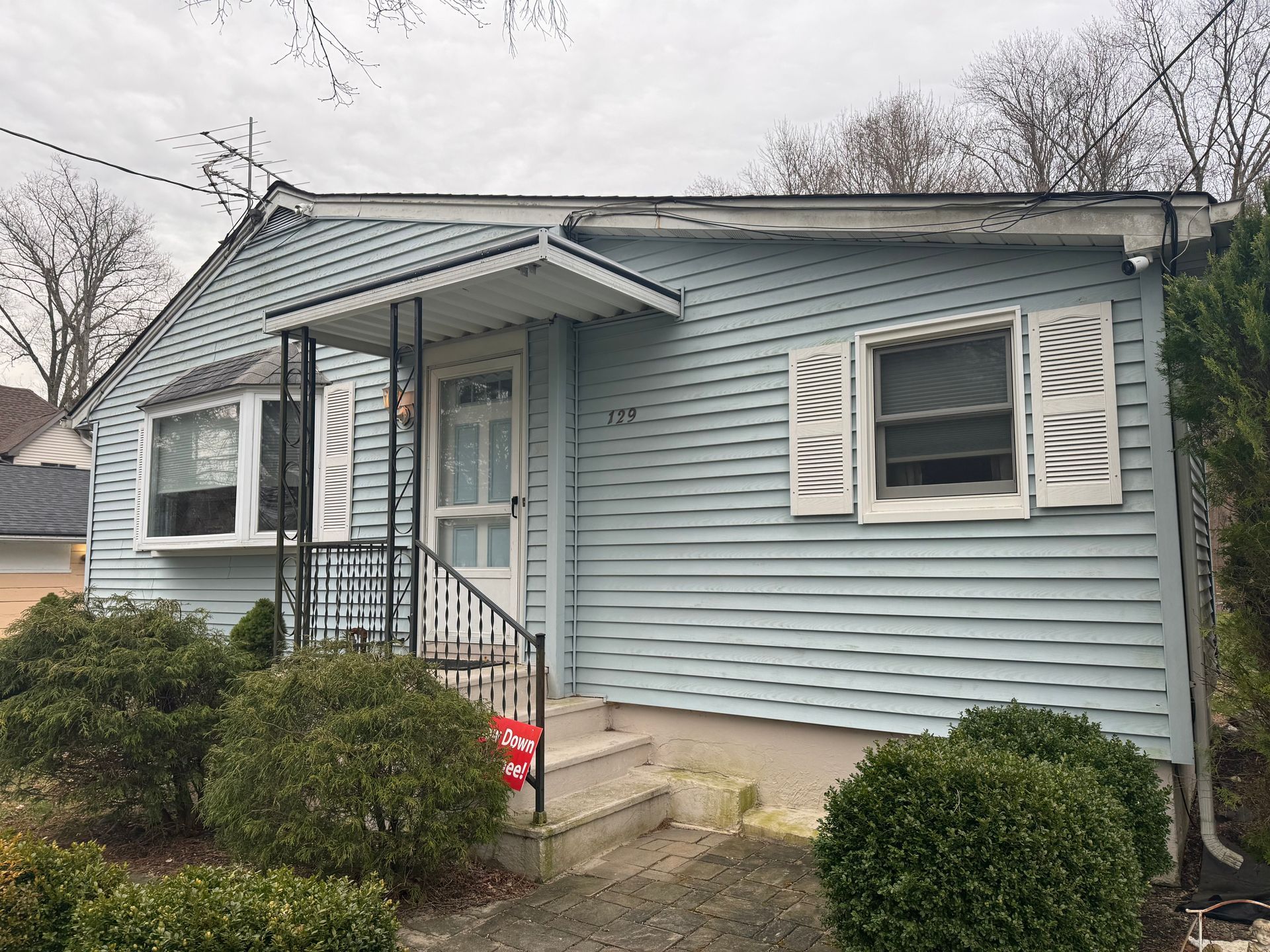 A small, single-story light blue house with horizontal siding, a covered porch, and a bay window under a cloudy sky.