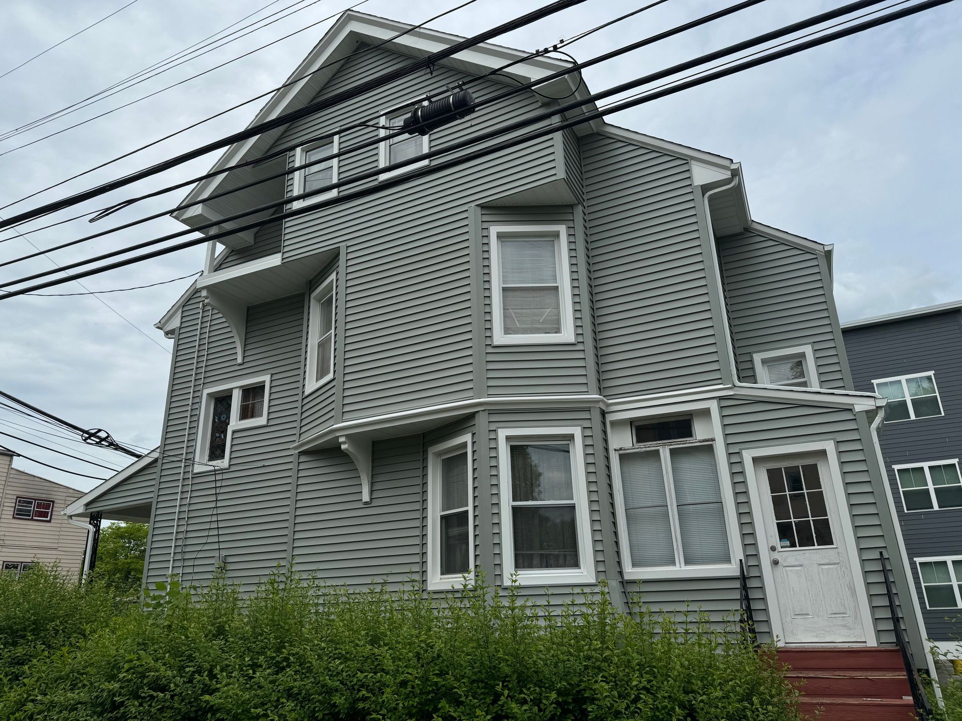 A gray, multi-story house with white trim, a gabled roof, and a green hedge in front under a cloudy sky.
