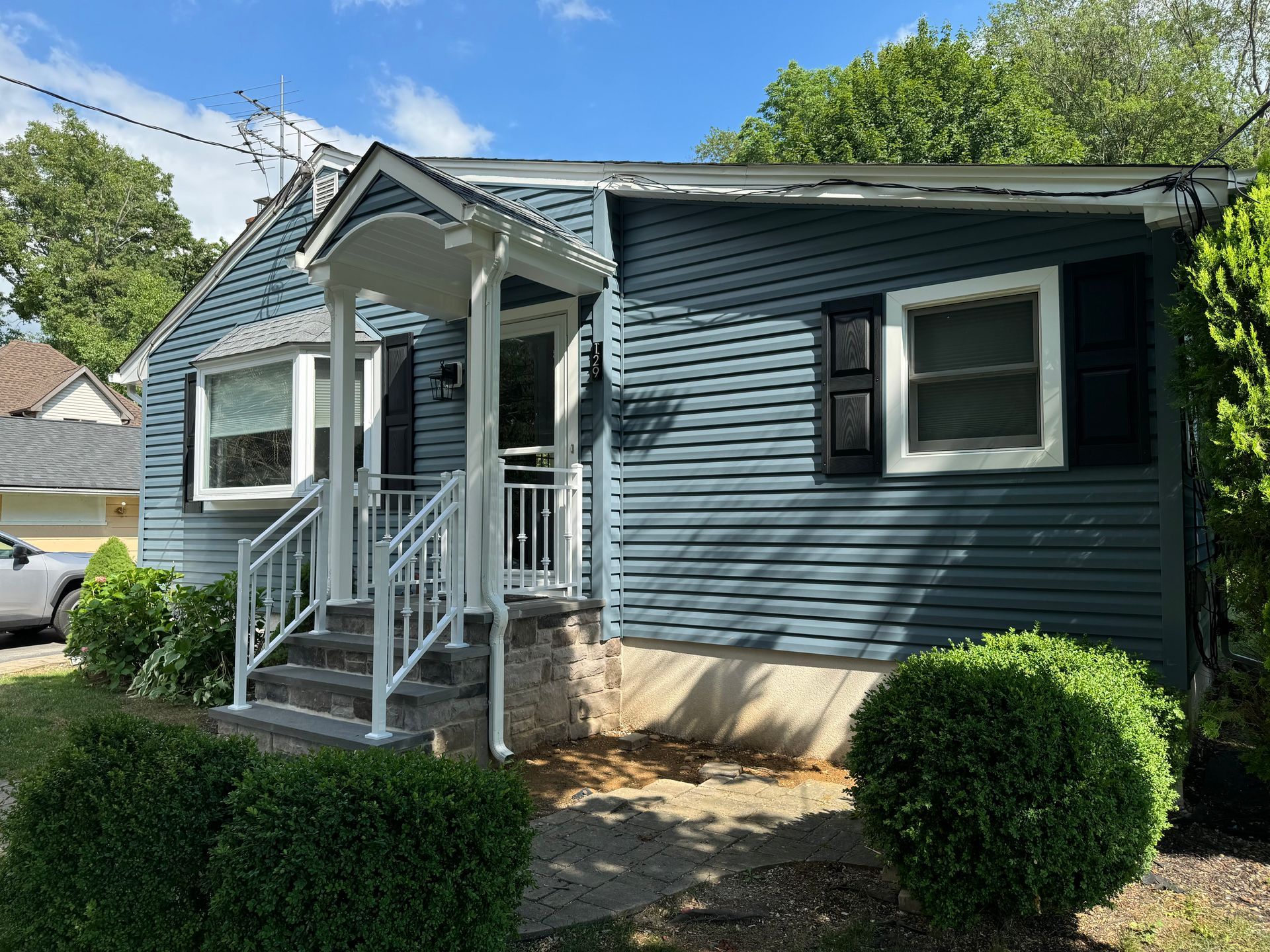 A blue-sided cottage with a white front porch, stairs with railings, and rounded bushes in the front yard.