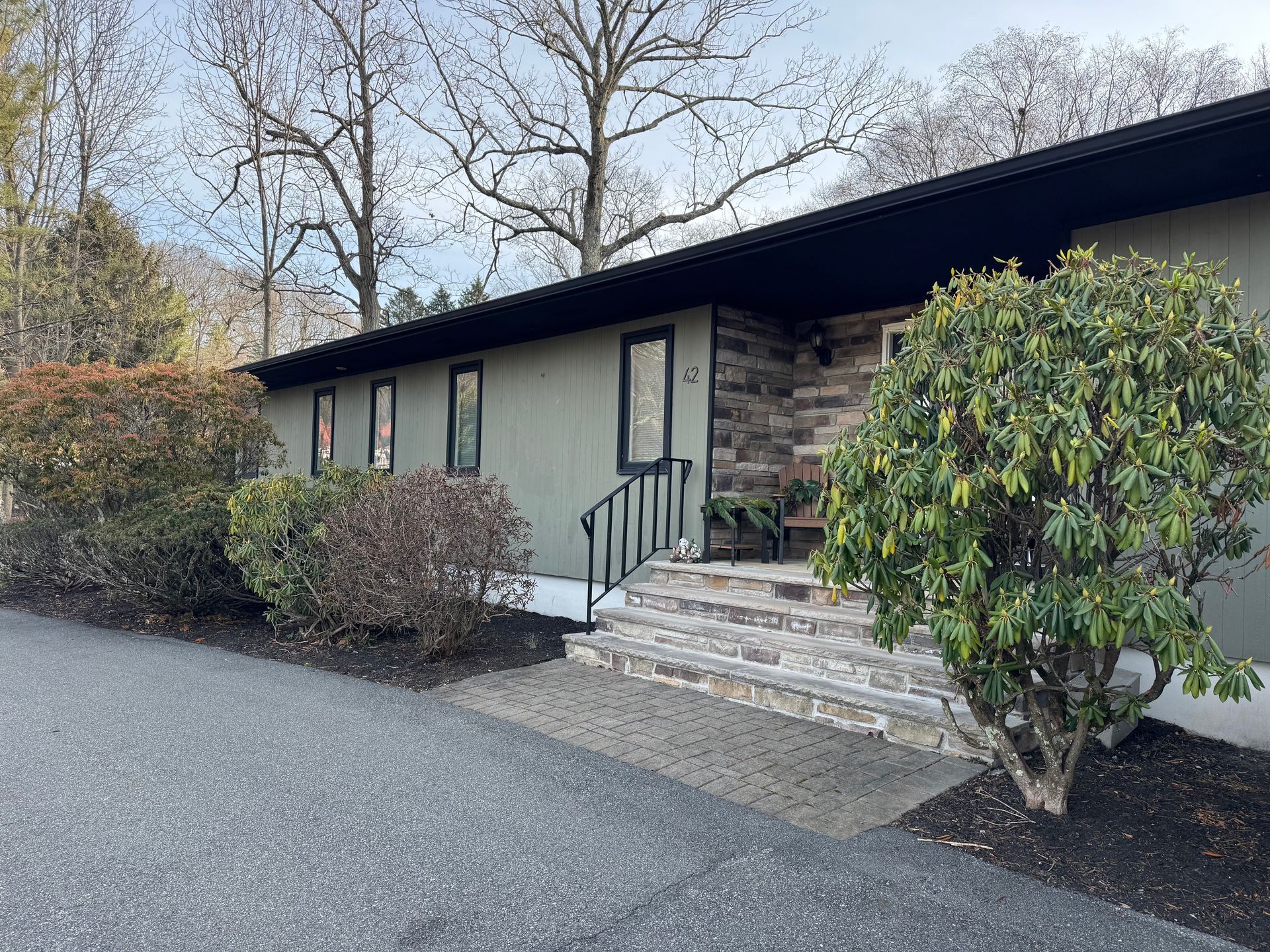 A tan, mid-century style ranch house with a dark roof and a stone-accented entryway, viewed from a gravel driveway.