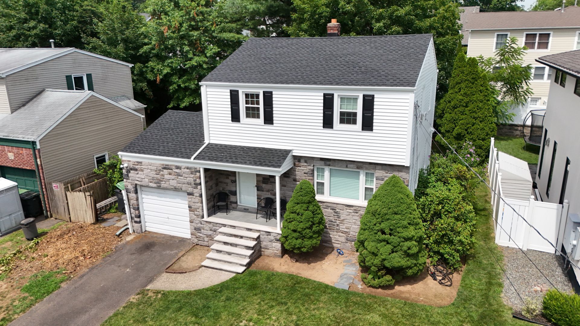A two-story house with white siding and a stone lower level, featuring a dark roof, front steps, and a small garage.