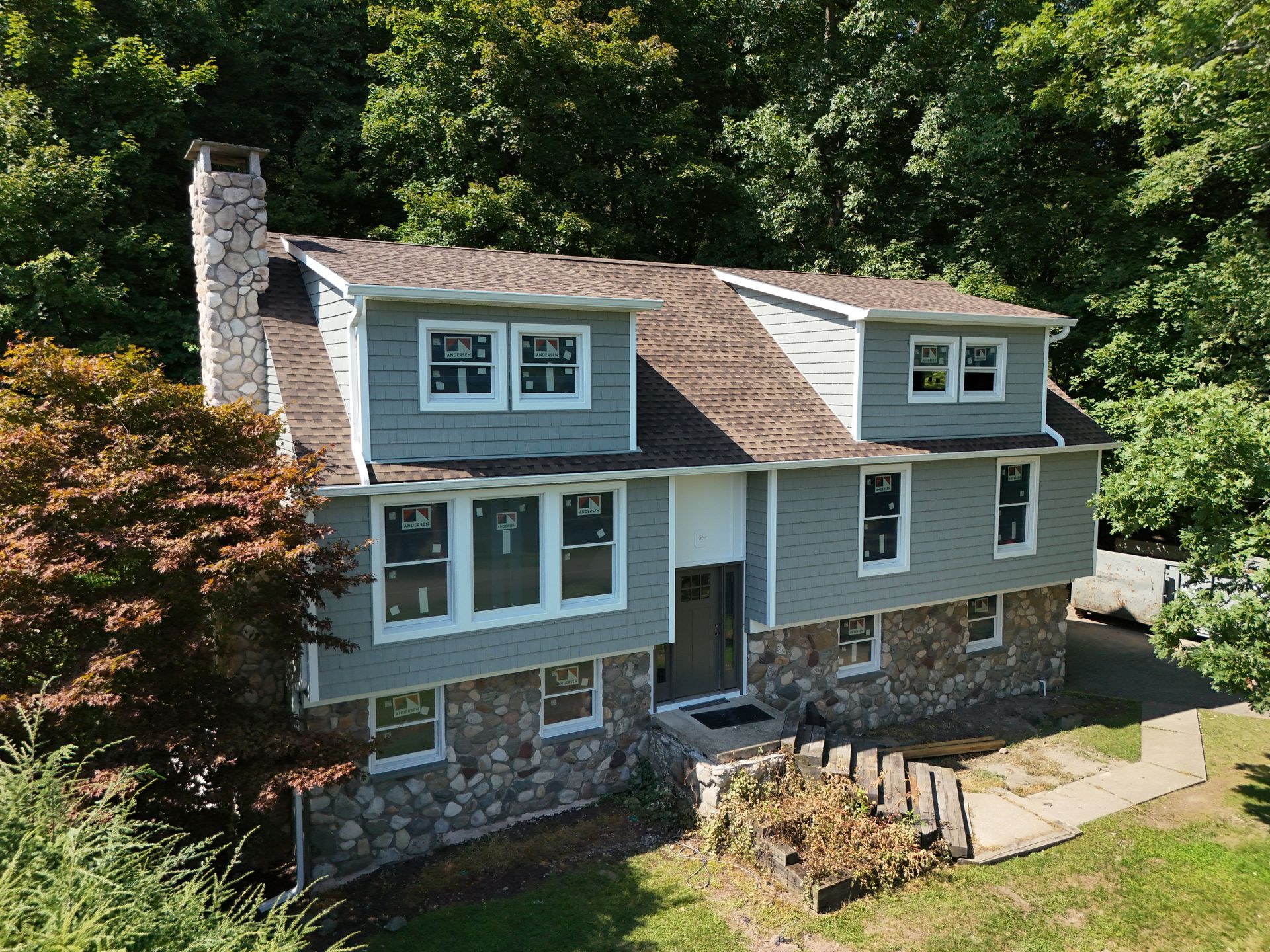 A light gray two-story house with a stone foundation, a matching stone chimney, and a brown shingled roof amid trees.