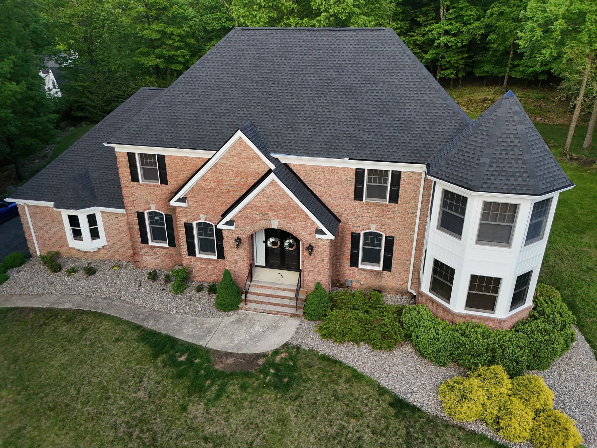 A high-angle view of a large, two-story brick home with a dark shingled roof, a turret, and stone landscaping.