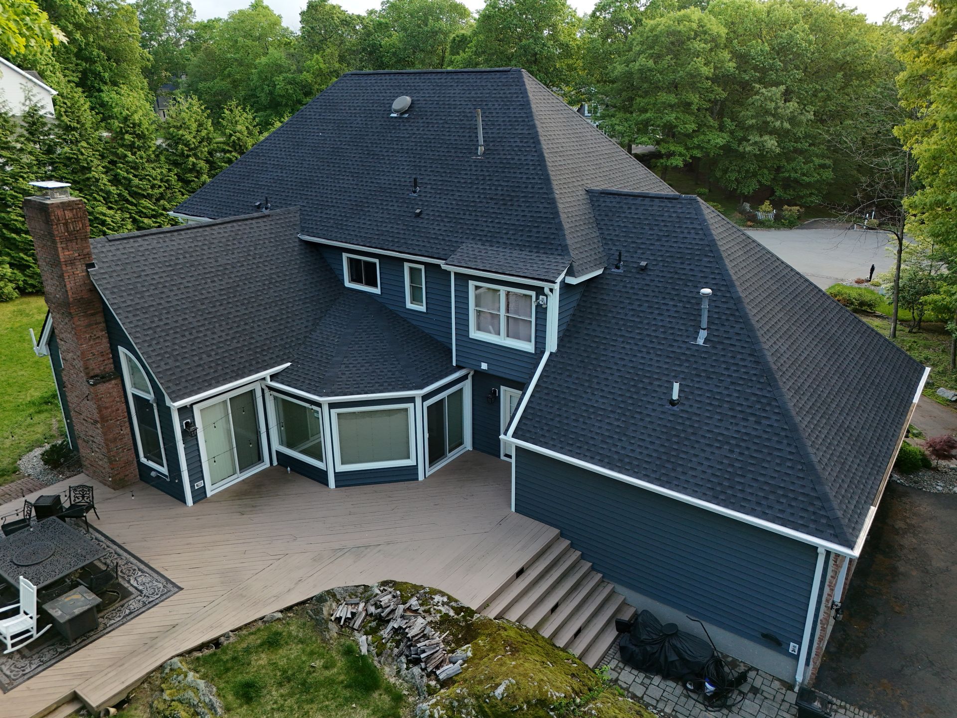 An aerial view of a dark blue two-story house with a black shingled roof, large windows, and a back patio.