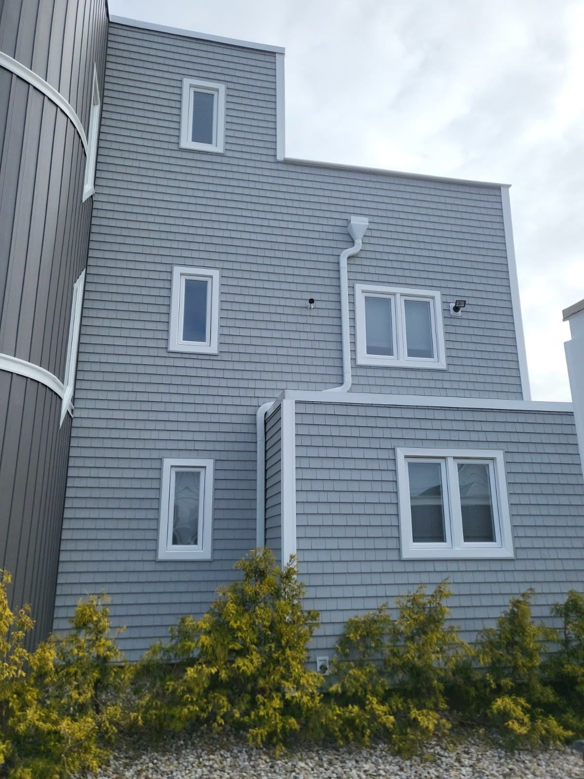 A light gray house with cedar shake siding, several vertical and rectangular white-trimmed windows, and small bushes.