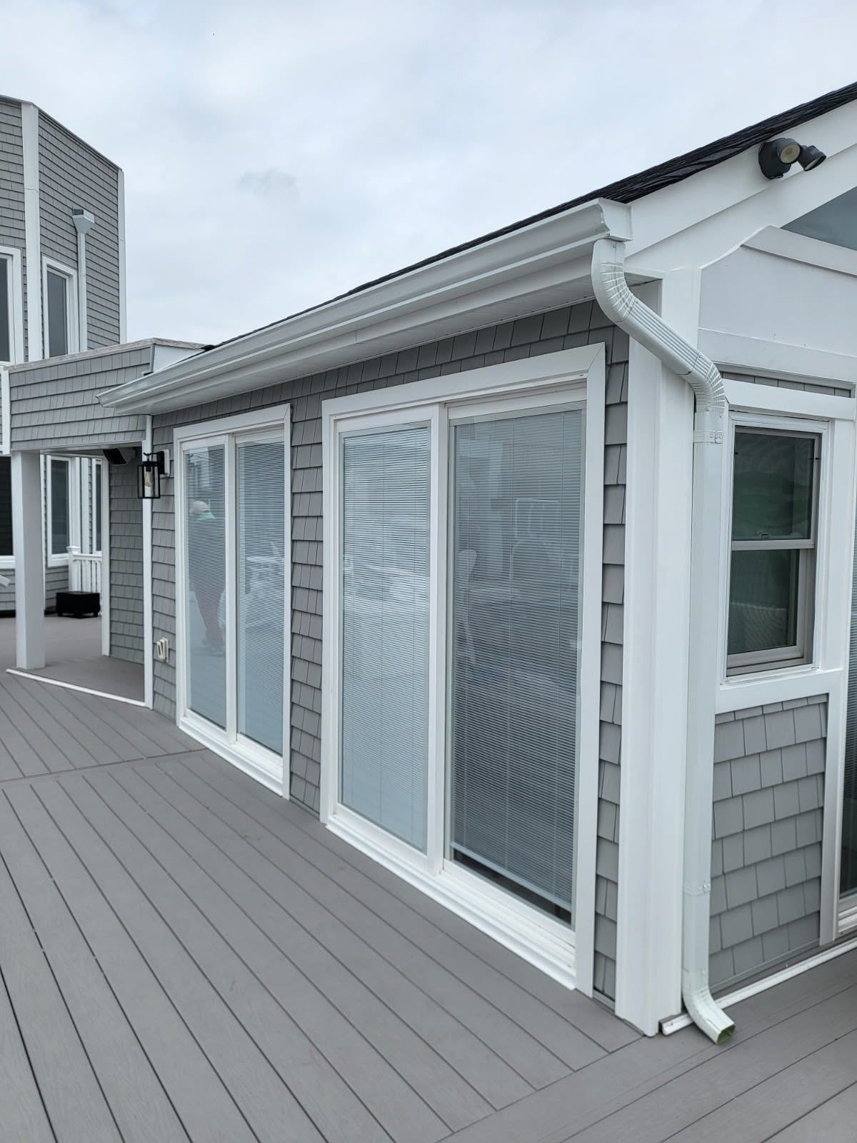 A grey shingled home exterior with sliding glass doors, white trim, and a deck under an overcast sky.
