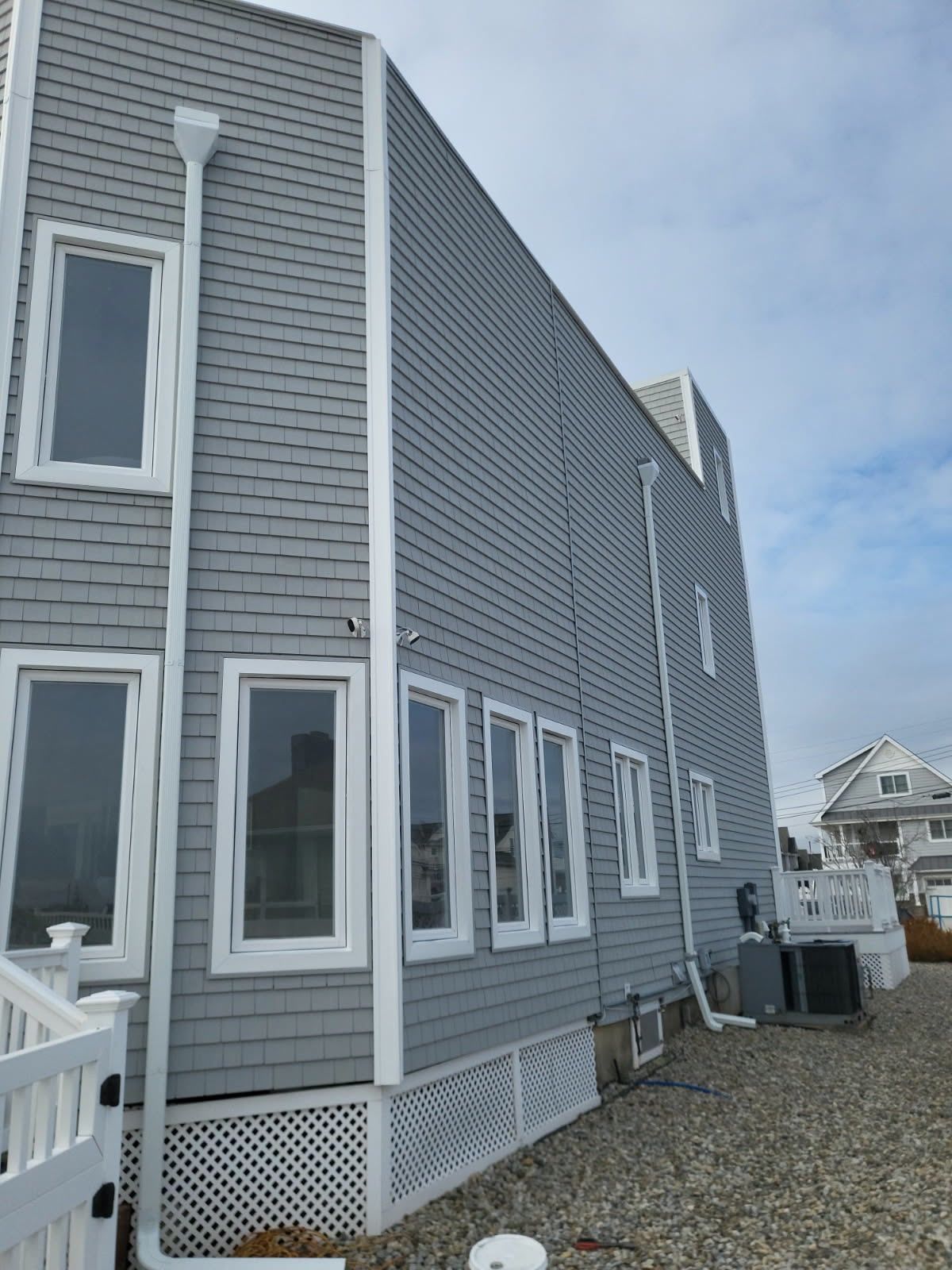 A side view of a gray-shingled two-story house with white trim, downspouts, and windows, situated on a gravel lot.