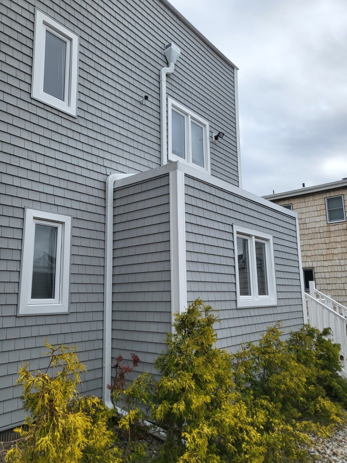 A grey shingled house exterior with white trim, two levels, white-framed windows, and small bushes in front.