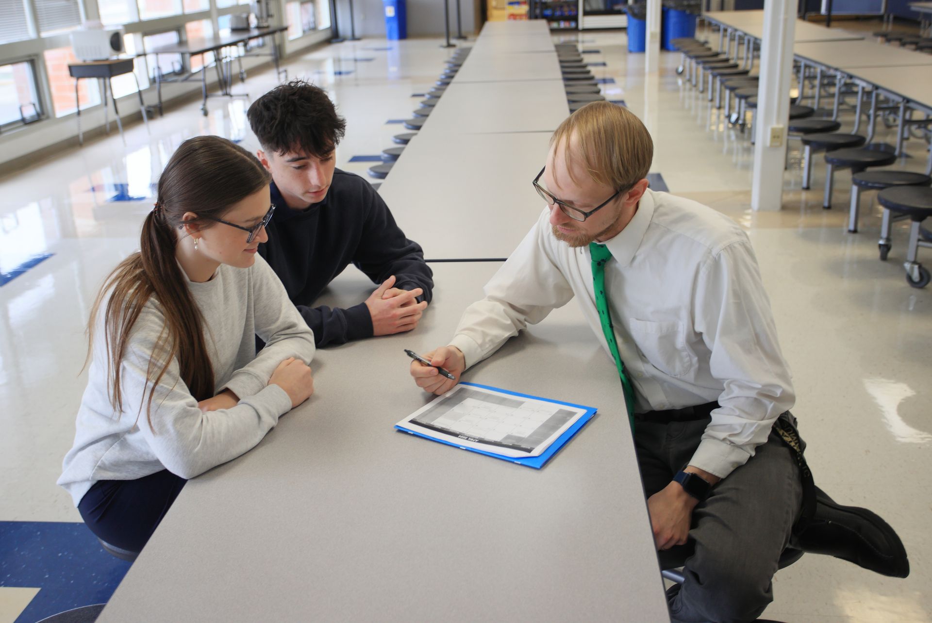 A man and two students are sitting at a long table in a cafeteria.