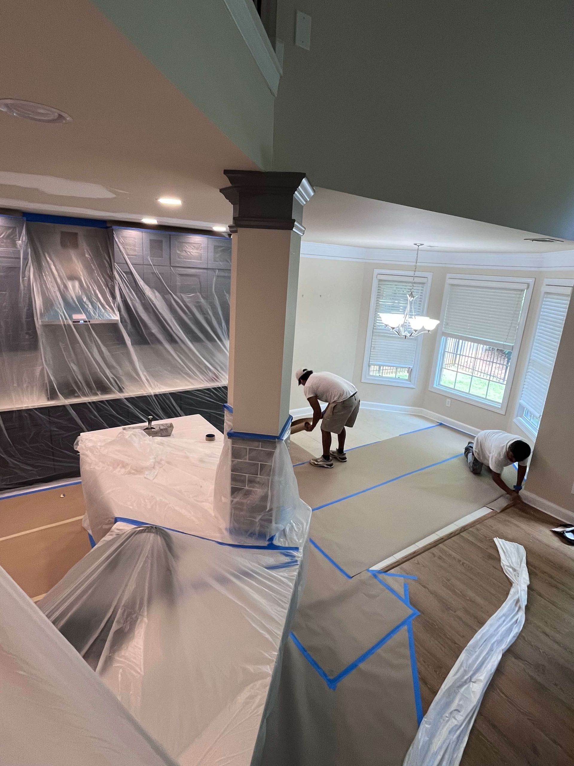 Two crew members preparing the dining area of a kitchen for painting, covering floors and furniture for protection.