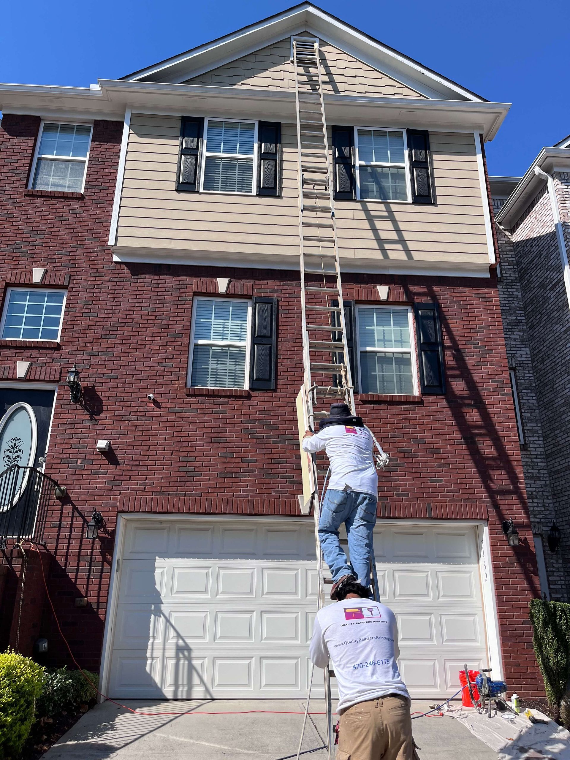 Professional painters using a ladder to safely access and paint the fascia, soffit, and gutters of a residential home.