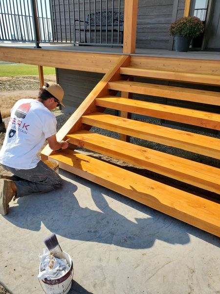 A man is kneeling down painting wooden stairs on a deck.