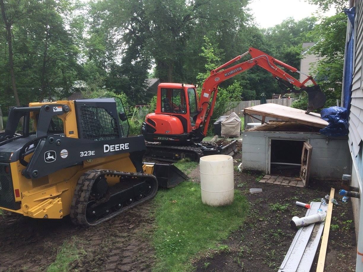 A small excavator and skid steer are used to demolish a small structure in a yard, with a house in the background.