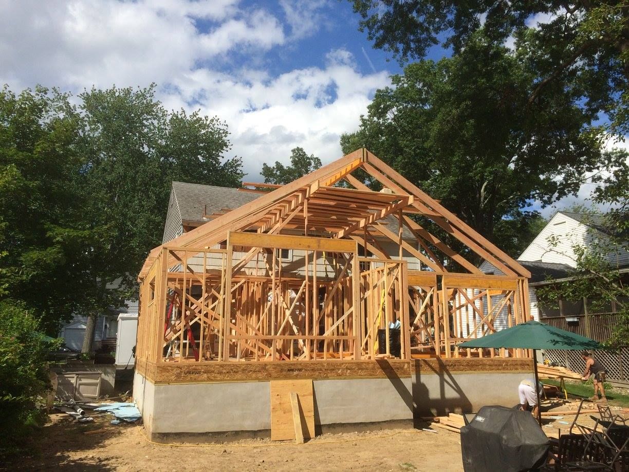 House under construction; wooden frame extension with roof supports, surrounded by trees.