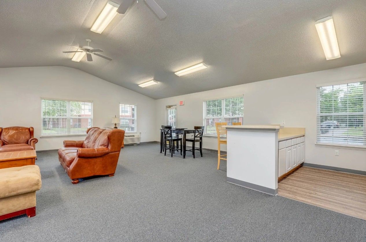 A bright, open-plan living room with grey carpeting, a brown leather sofa, dining table, and a kitchen island.