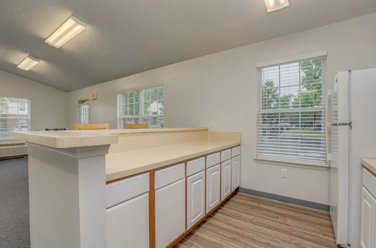 A bright kitchen area with white cabinets, a light-colored countertop, and a window with blinds beside a refrigerator.