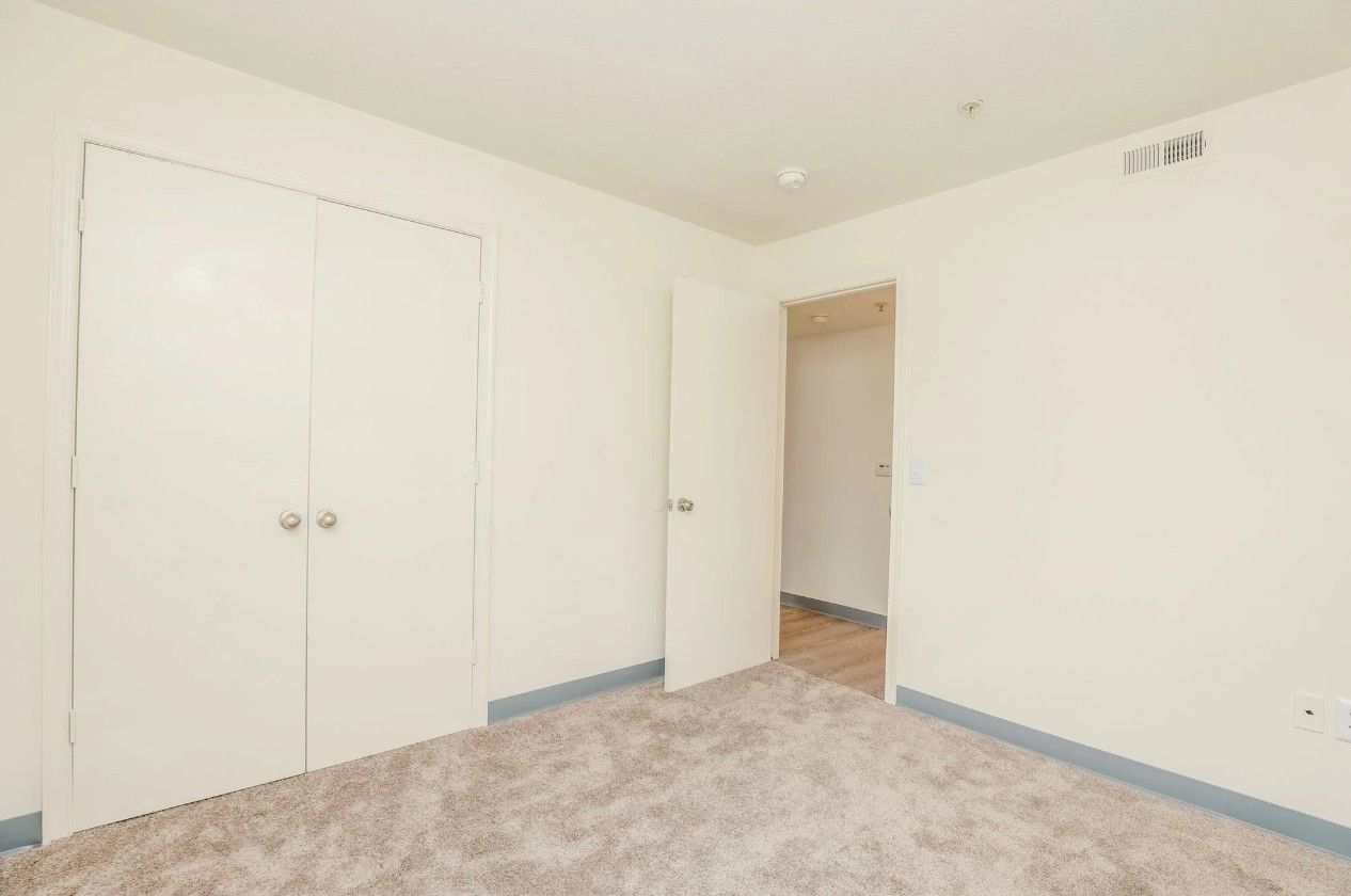An empty room with white walls, gray carpet, a double-door closet, and an open doorway leading to another room.
