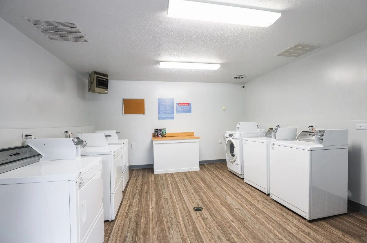 A laundry room with multiple white washers and dryers arranged along the walls on a light wood-look floor.