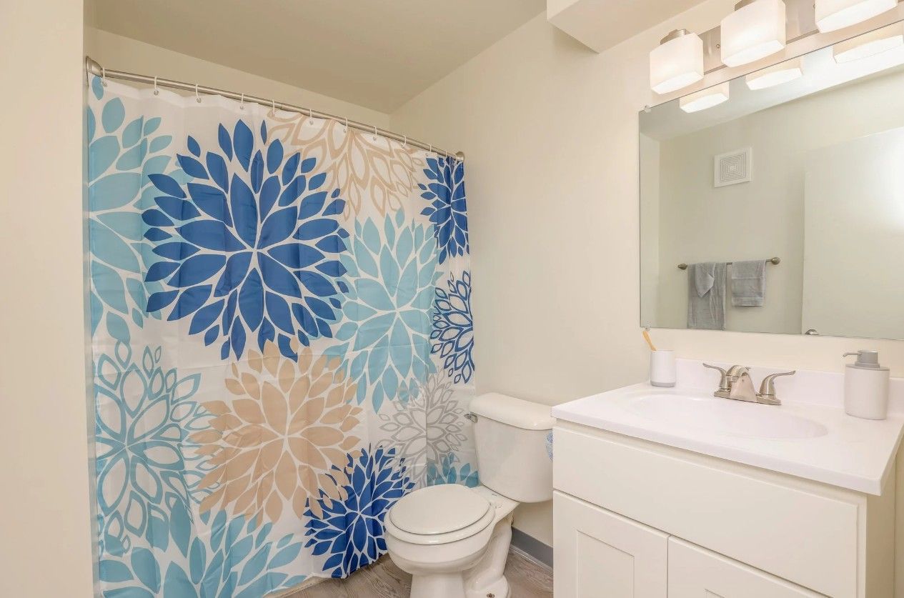 A bathroom with a shower curtain featuring blue and beige flower designs, a toilet, and a white vanity with a mirror.