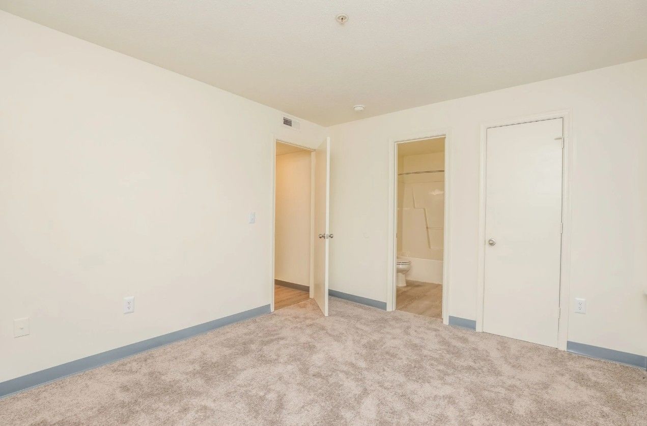 A vacant room with light-colored walls and carpet, featuring two doorways leading to a hallway and a bathroom.