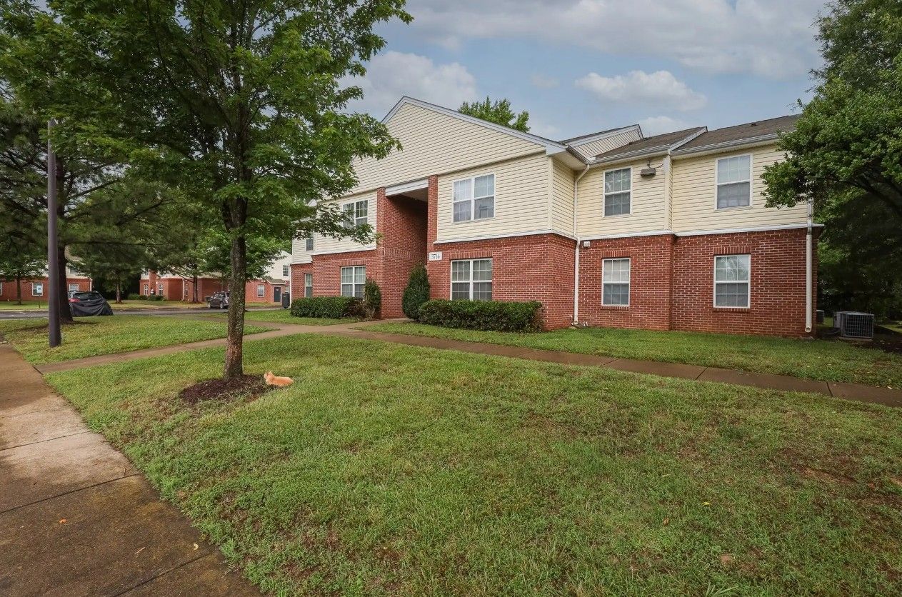 A two-story apartment building with beige siding and brick ground-level walls, surrounded by trees and a grass lawn.