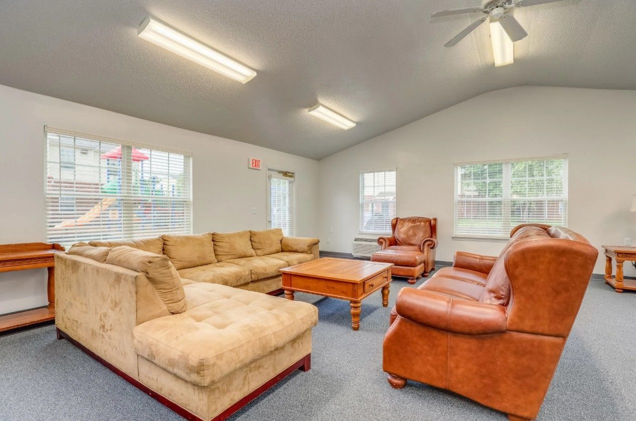 A furnished lounge featuring a large tan sectional sofa, a brown leather armchair, a wooden coffee table, and large windows.