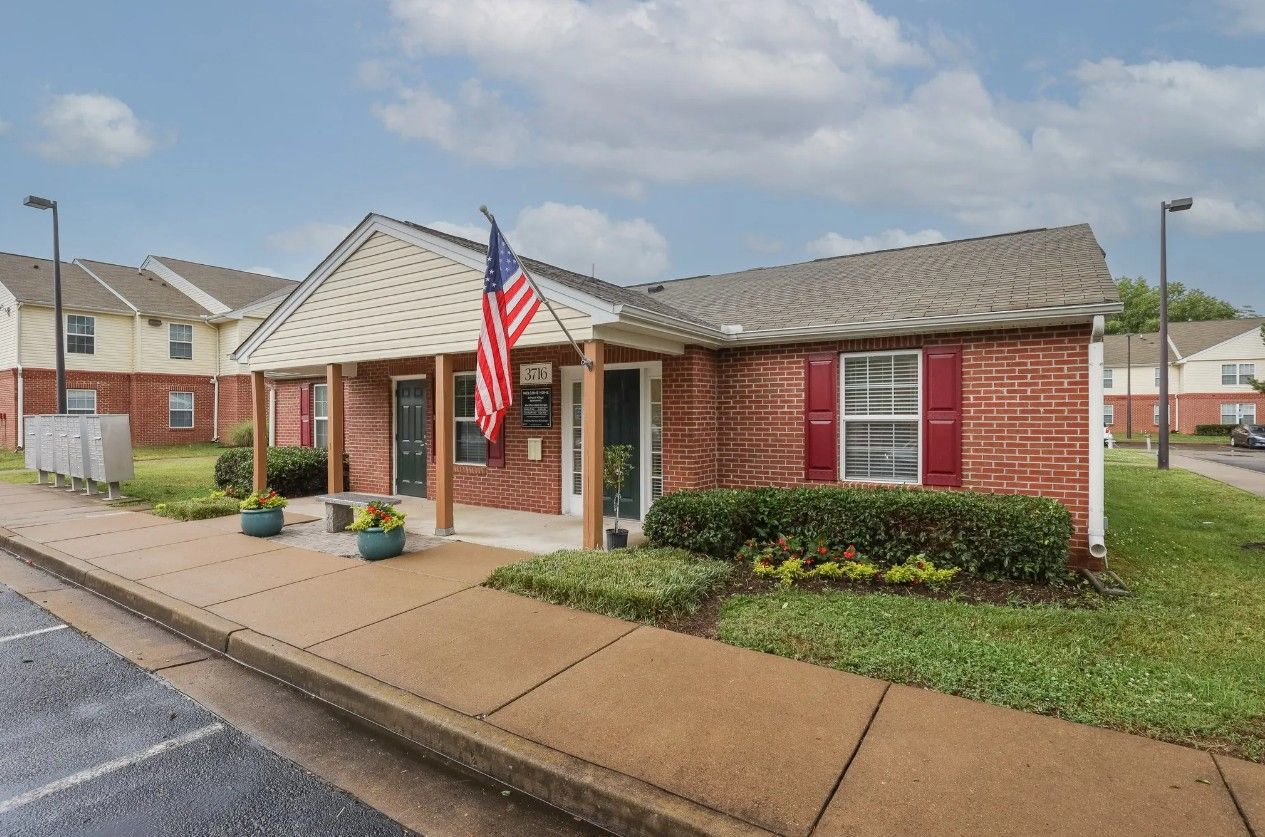A one-story brick leasing office building with a porch, American flag, and surrounding landscaping.